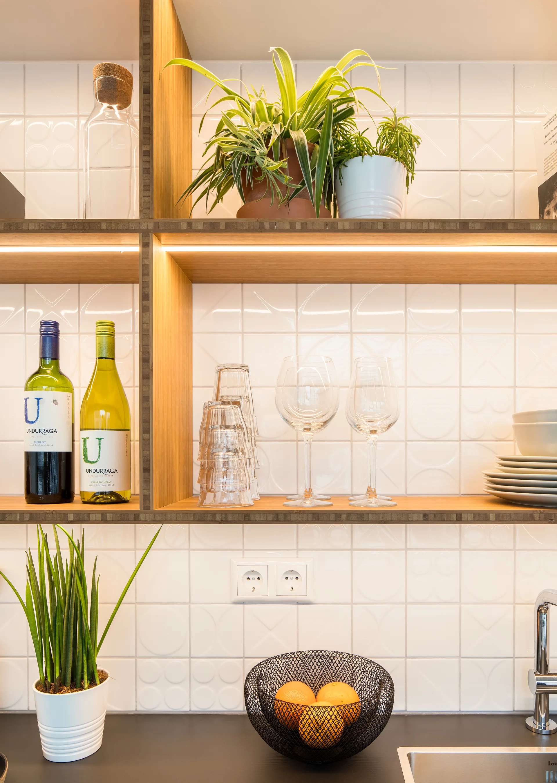 Open wood kitchen shelves with integrated LED strip lights display glassware and plants against a white patterned tile backsplash.
