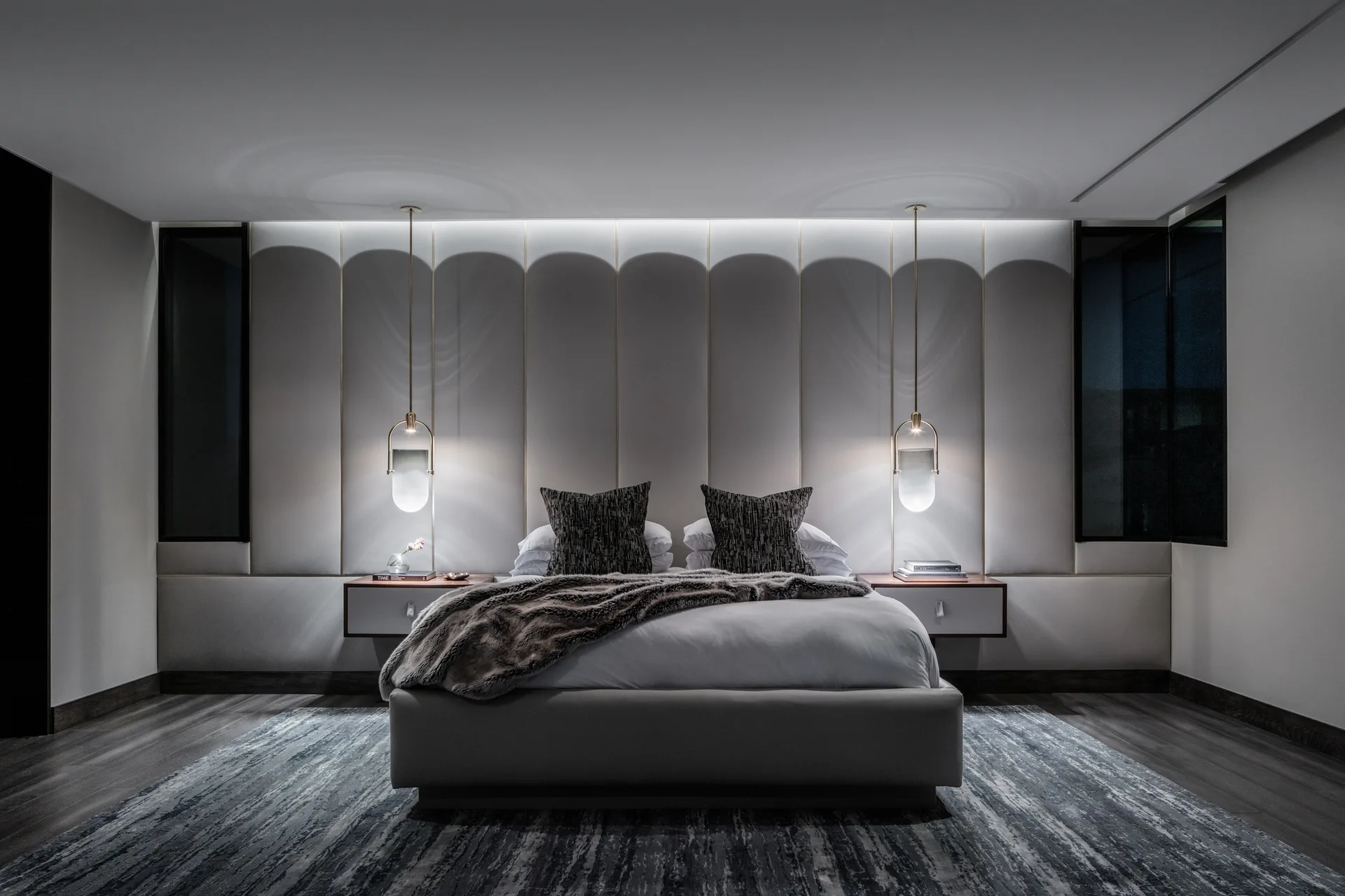 A contemporary gray bedroom with a platform bed, vertical upholstered wall panels, hanging brass pendant lights, and floating nightstands.