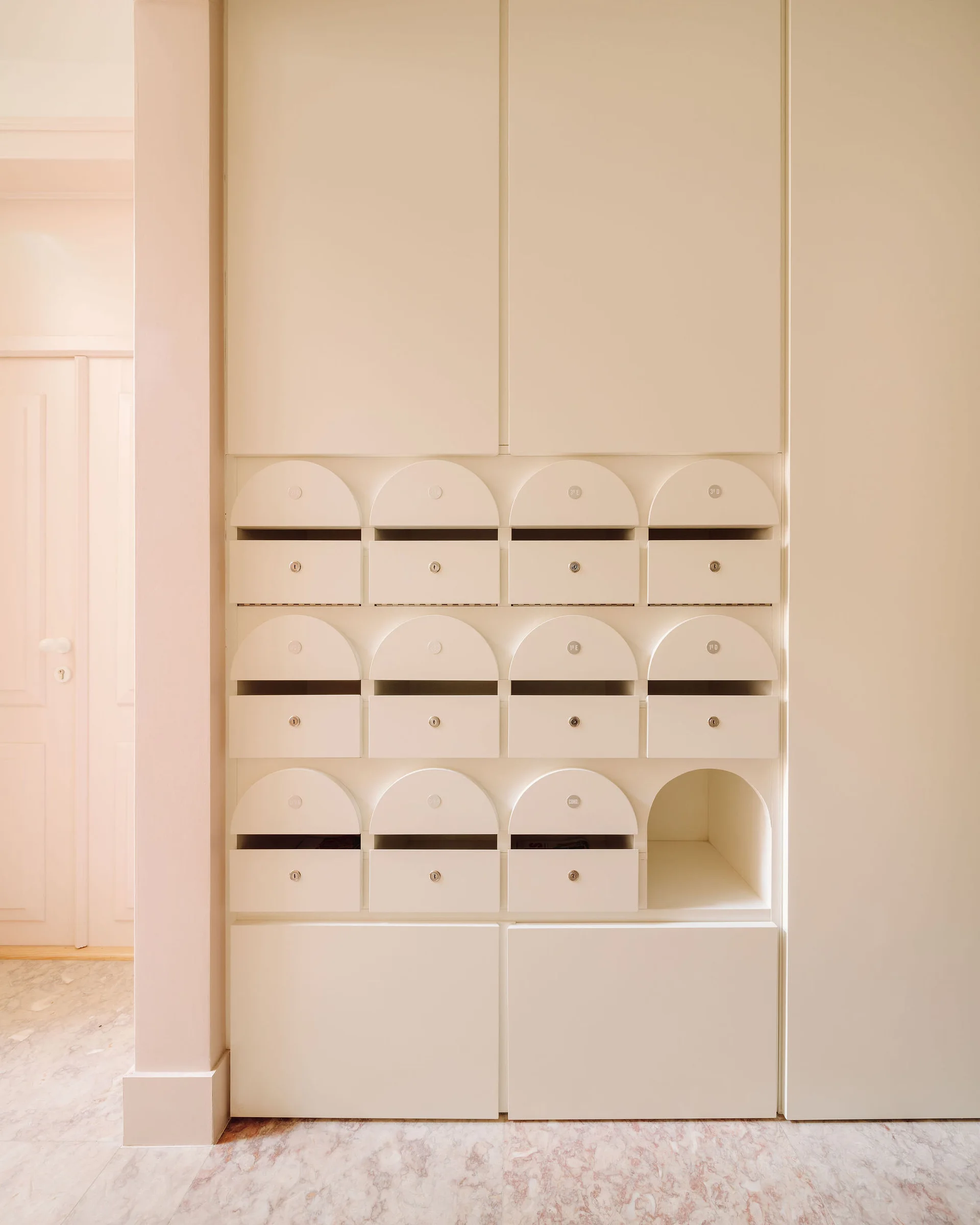 Cream-colored built-in entryway cabinetry featuring a grid of arched mailboxes with key locks above a veined marble tile floor.