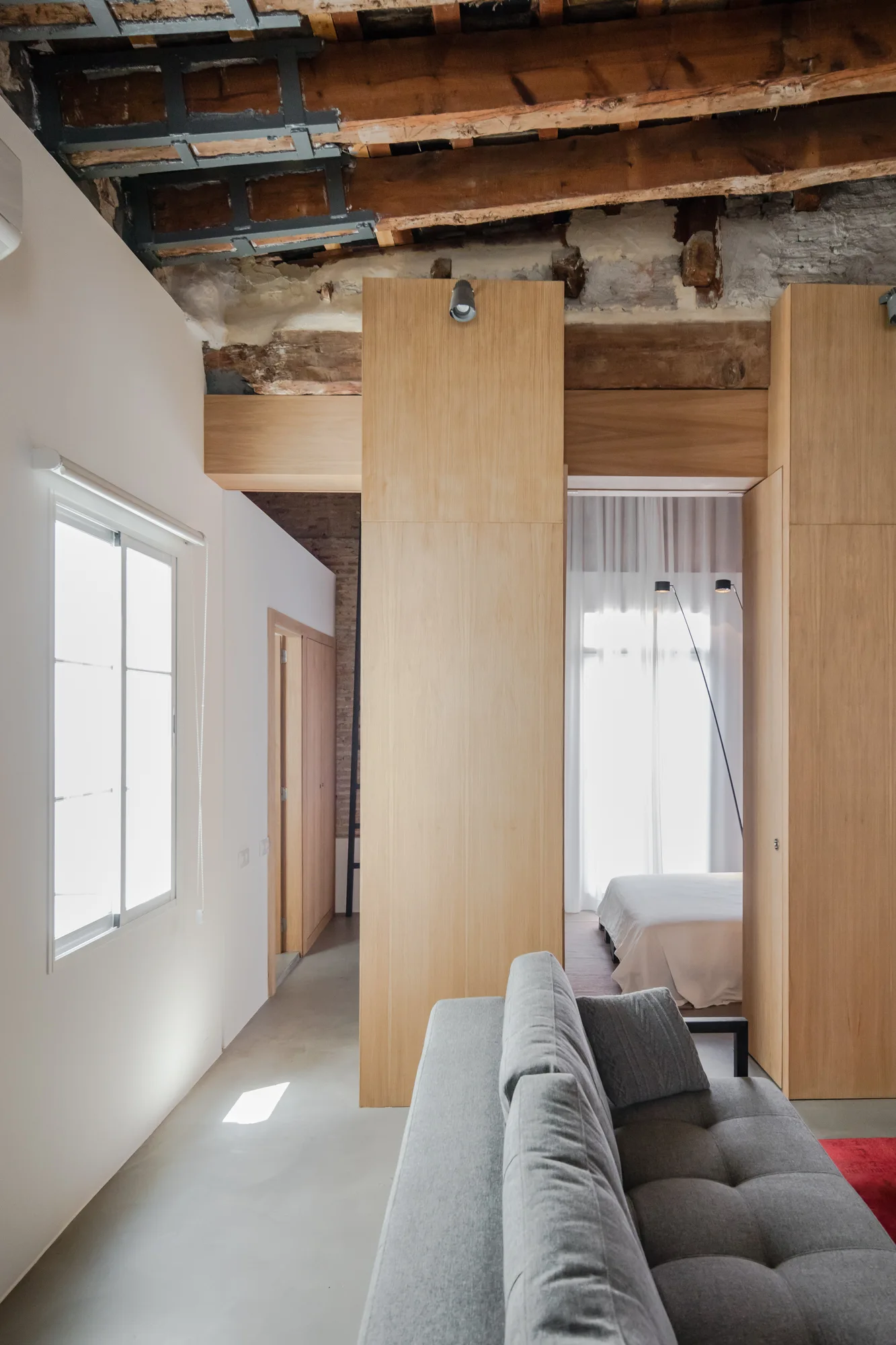 A grey tufted sofa sits in a modern apartment featuring light wood partition walls and rustic exposed timber ceiling beams.