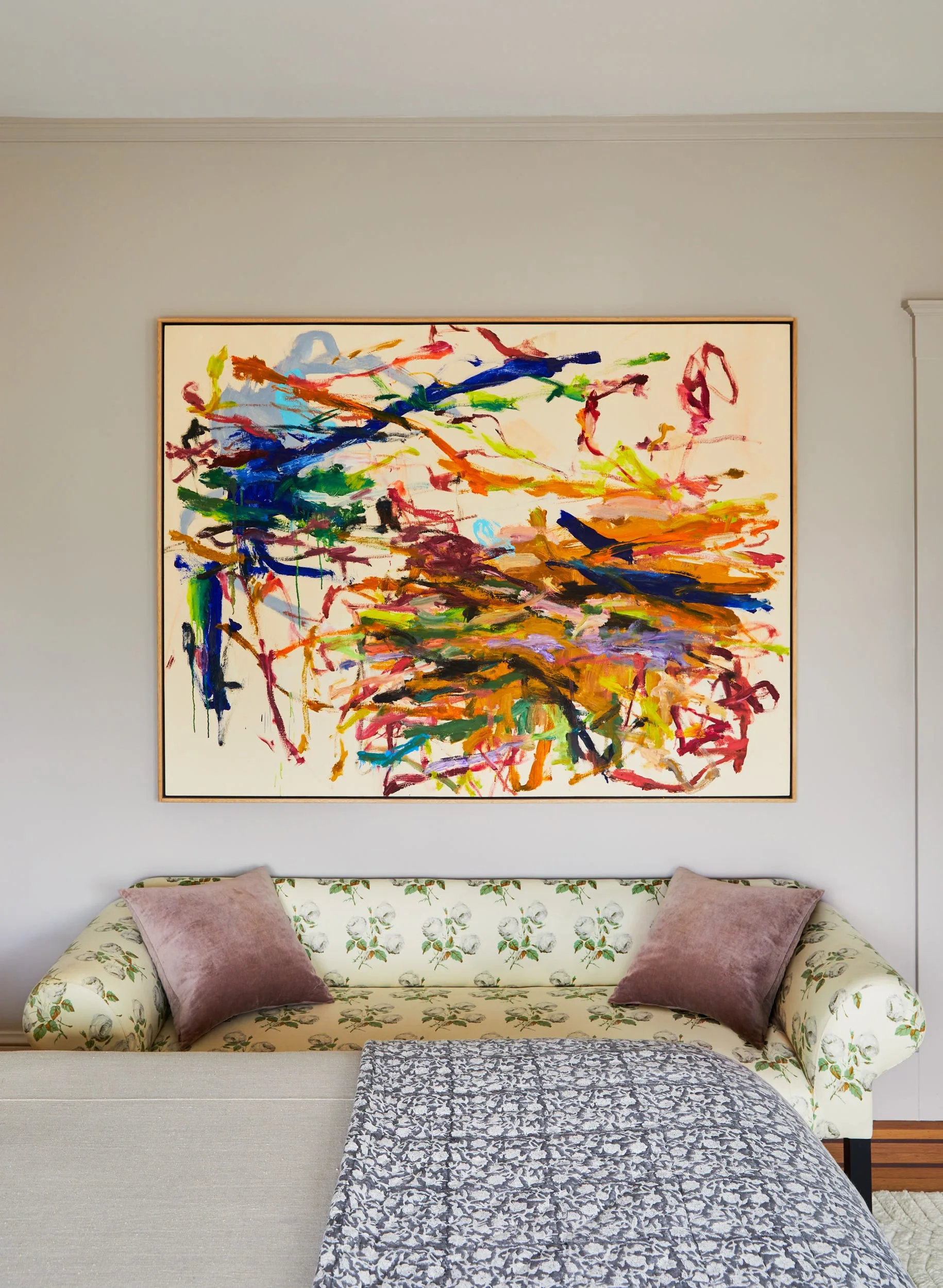 A large colorful abstract painting hangs on a light gray wall above a floral-patterned loveseat styled with mauve velvet throw pillows.