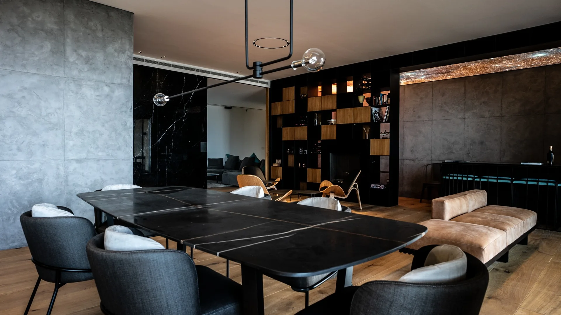 A modern dining room features a long black table surrounded by dark grey chairs, set against a concrete wall and a black geometric shelving unit.