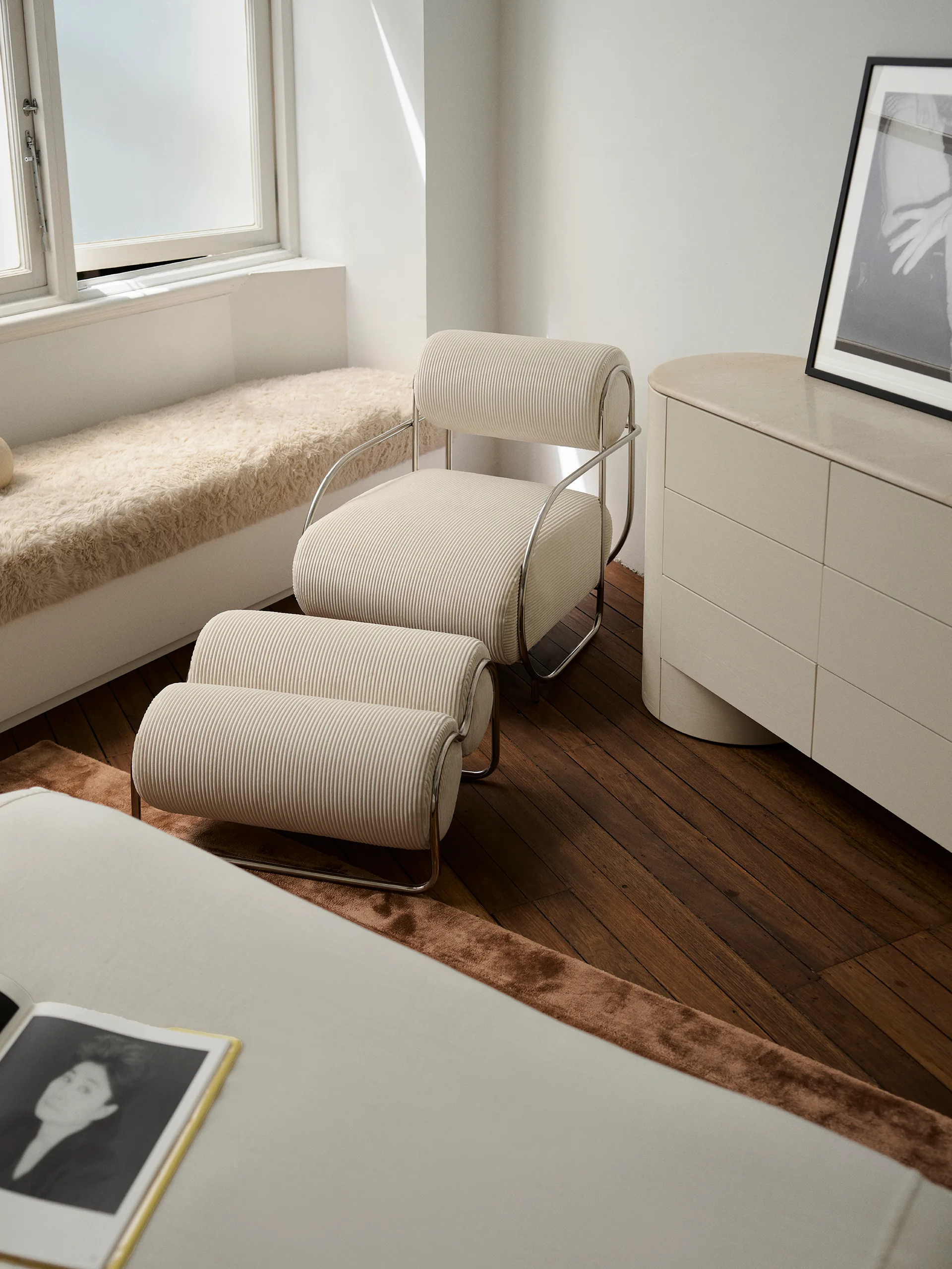 A neutral-toned bedroom features a ribbed cream armchair and matching ottoman with chrome frames next to a curved dresser on dark wood floors.
