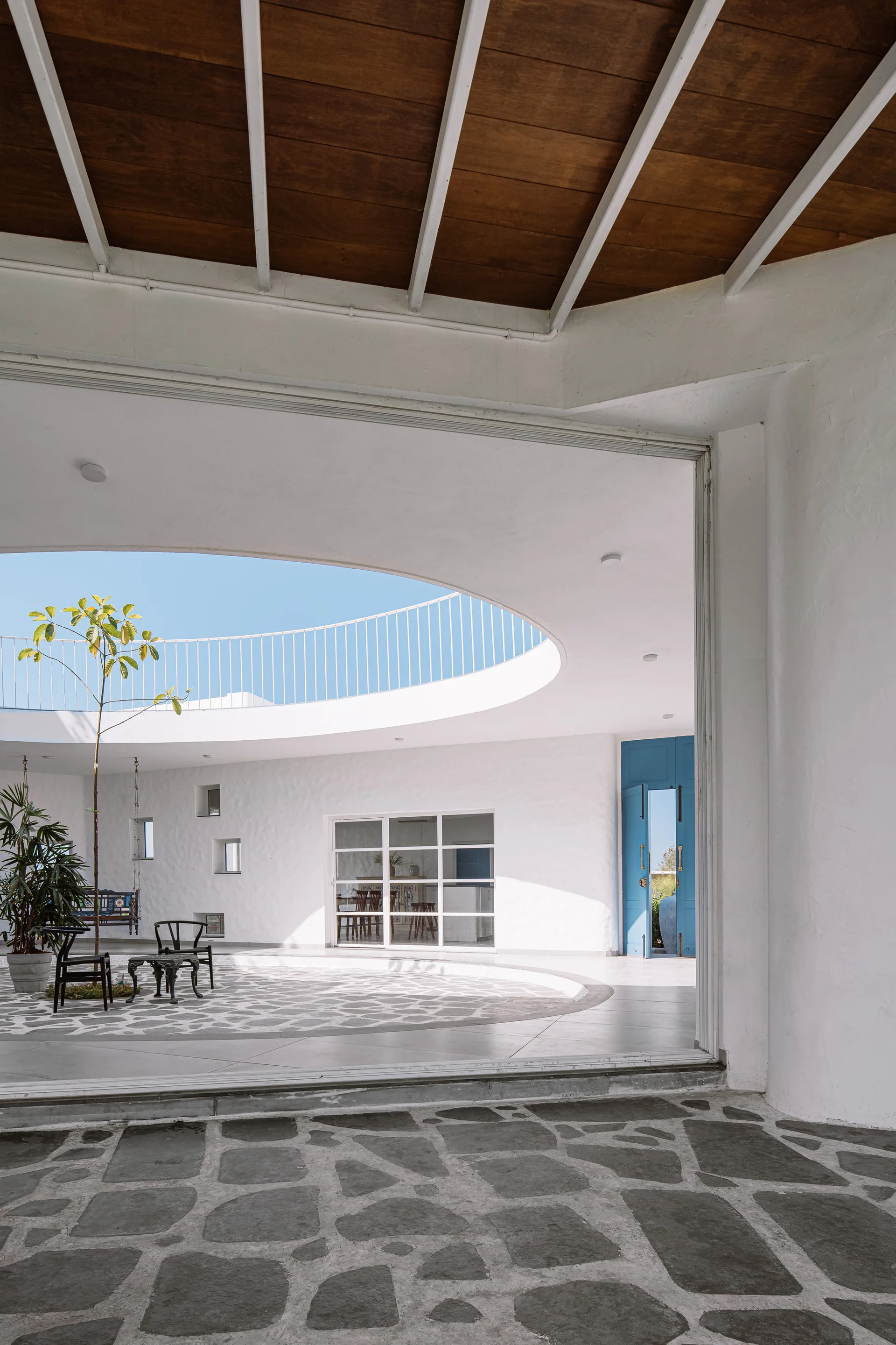 A white stucco courtyard features a large circular roof opening, irregular grey stone paving, and a vibrant blue double door.