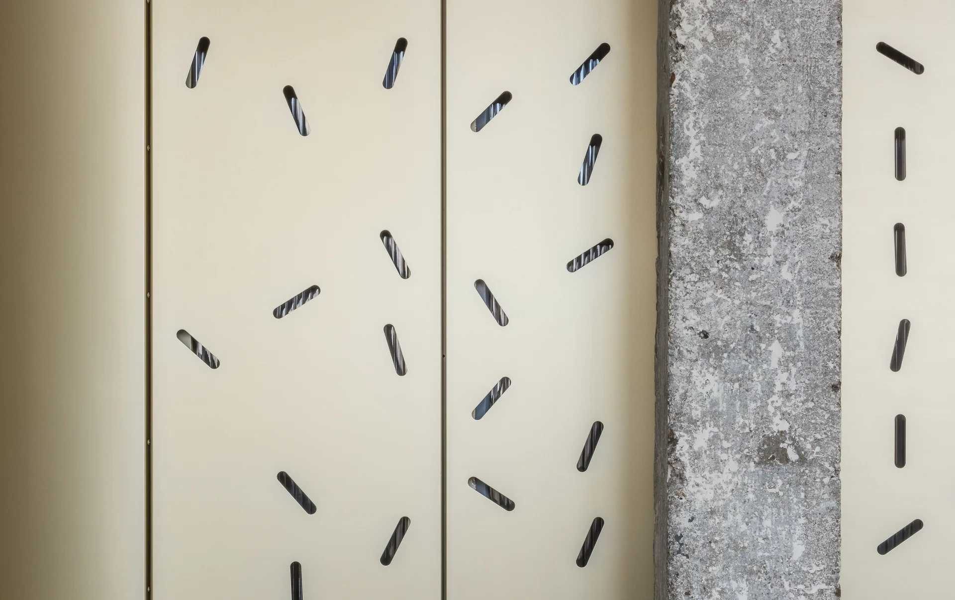 Off-white metal wall panels featuring diagonal slotted perforations next to a rough grey concrete column.