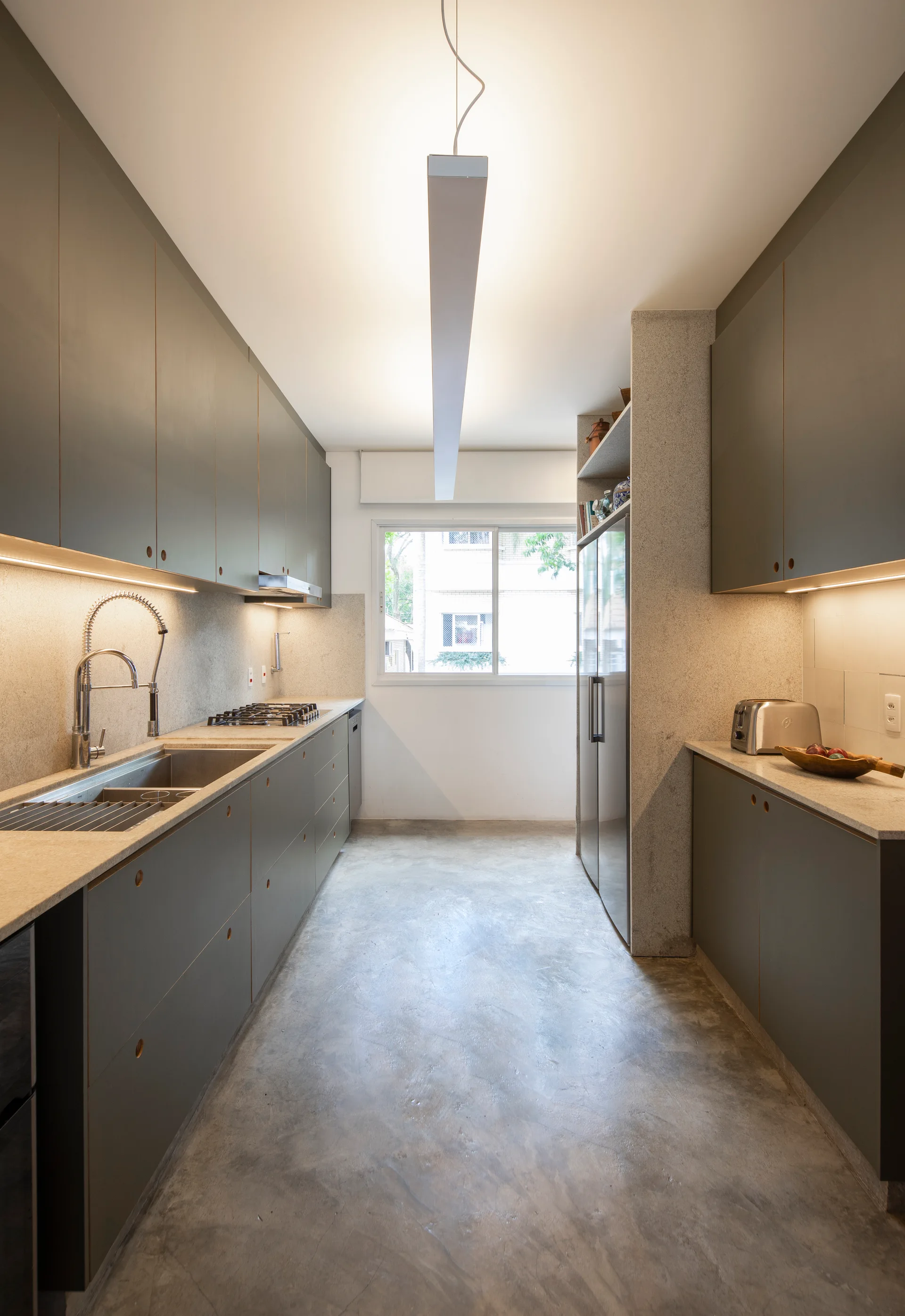 A minimalist galley kitchen features matte grey slab cabinets, polished concrete floors, and a long linear light fixture hanging from the ceiling.
