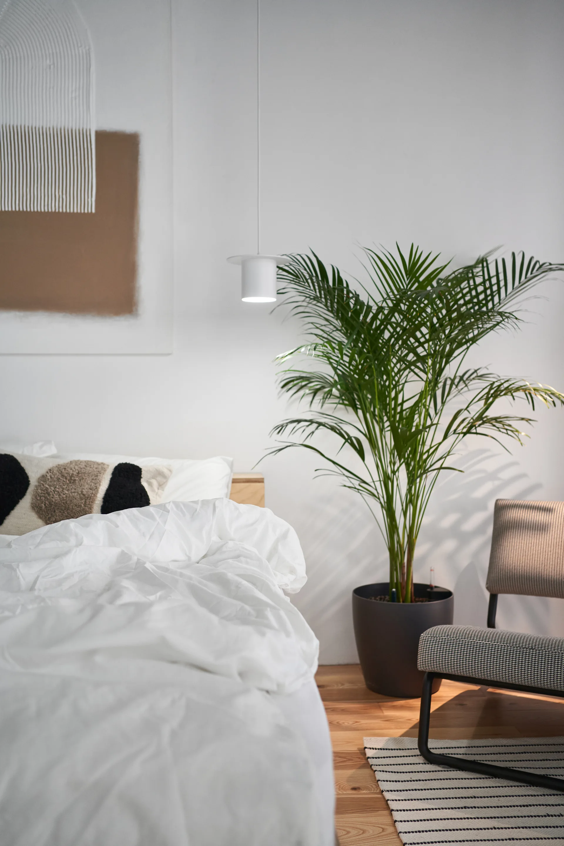 A minimalist bedroom featuring a bed with white linens, a tall potted palm, a houndstooth accent chair, and a hanging white pendant light.