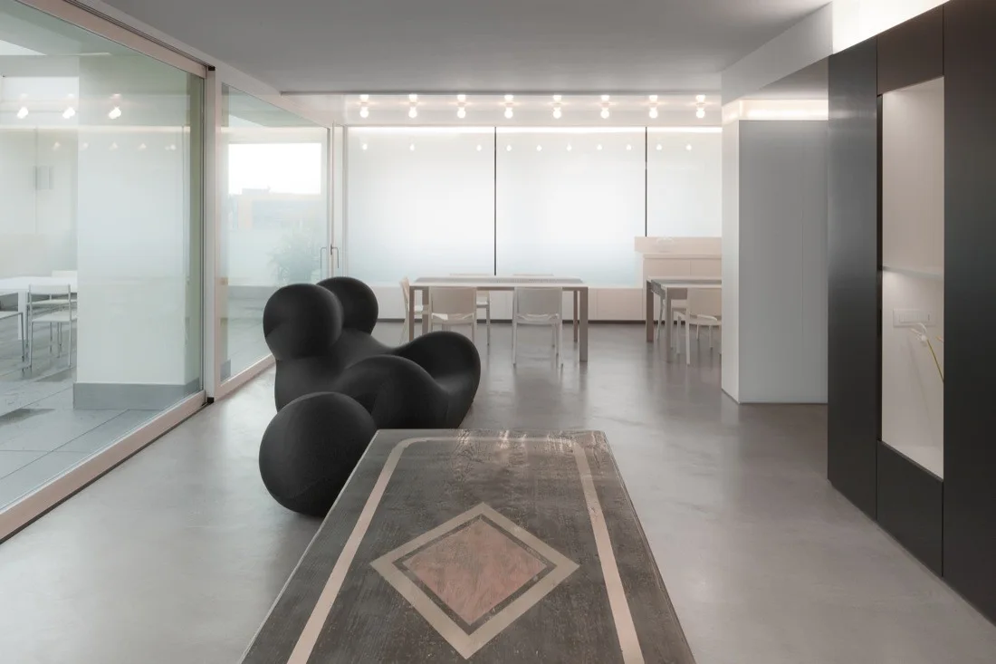 A minimalist grey living room featuring a bulbous black sculptural lounge chair, a wooden table with a diamond inlay, and sliding glass doors.