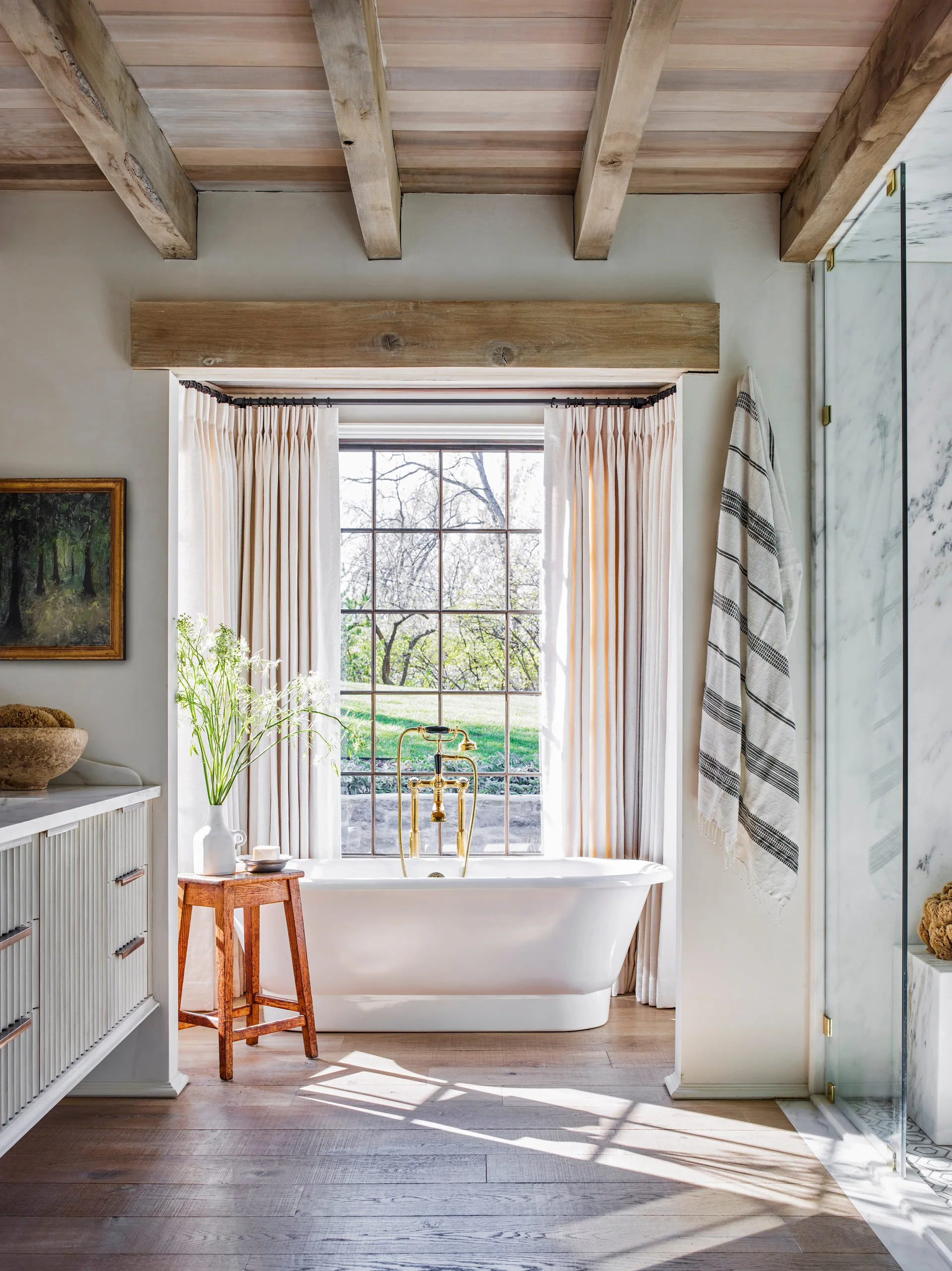 A bright bathroom features a white freestanding bathtub beneath a grid window, a fluted vanity, and exposed wooden ceiling beams.