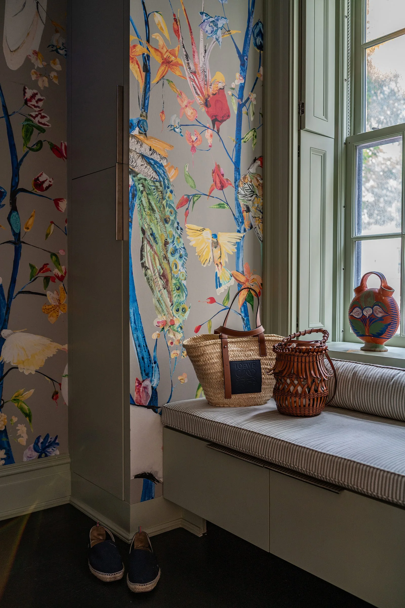 An entryway nook features a built-in window seat with a pinstripe cushion, sage green trim, and vibrant bird and botanical wallpaper.