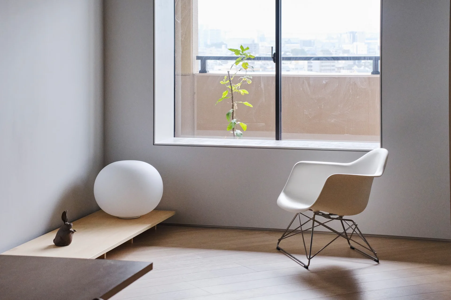 A minimalist room corner features a white molded armchair, a spherical floor lamp on a low wooden bench, and a large window.