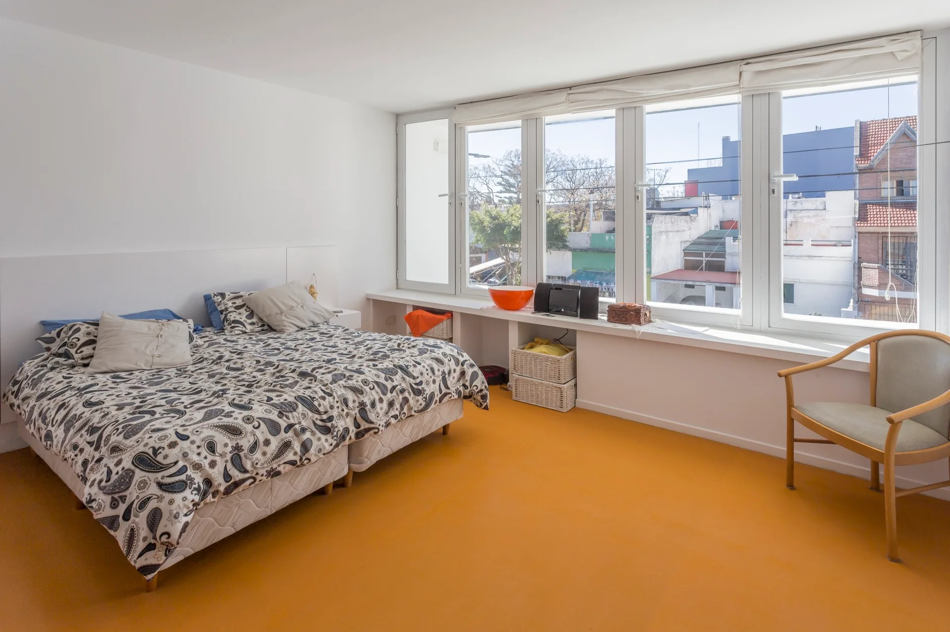 A minimalist bedroom featuring a bed with patterned paisley bedding, a bright orange floor, and a long white built-in ledge under a row of windows.
