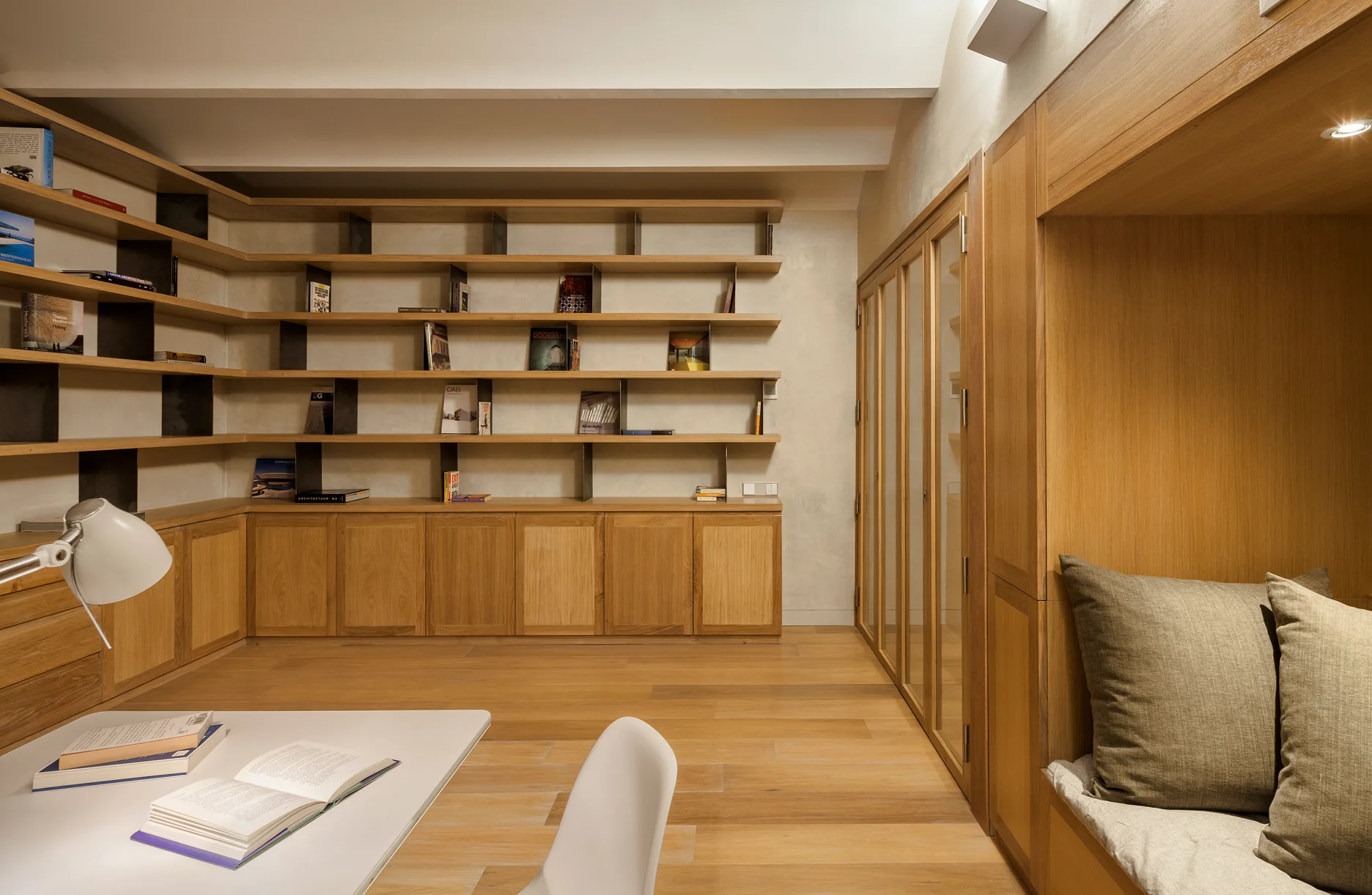 A minimalist home office featuring light oak built-in bookshelves, a corner reading nook, a white desk, and natural wood flooring.