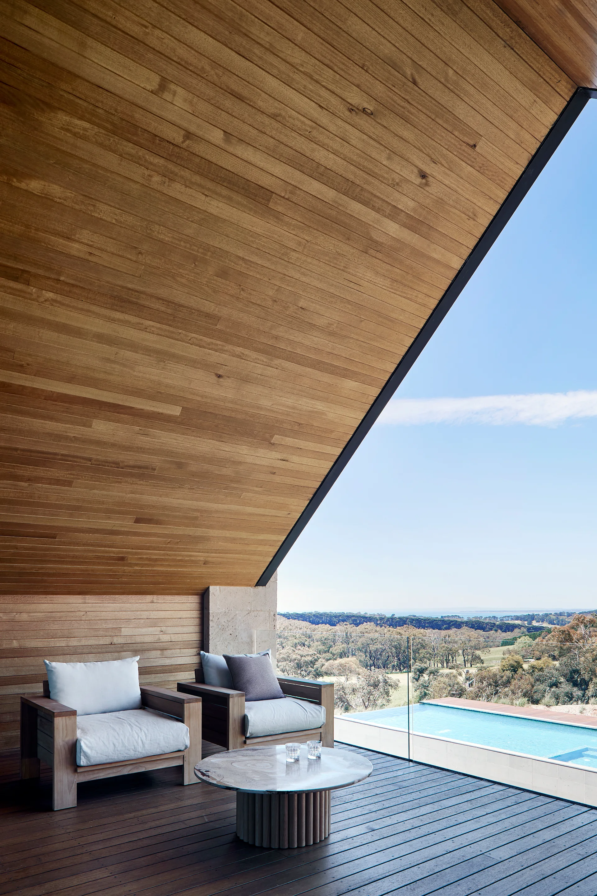 Two modern wood armchairs with white cushions and a stone coffee table sit on a dark wood terrace under a sloped timber ceiling overlooking a pool.