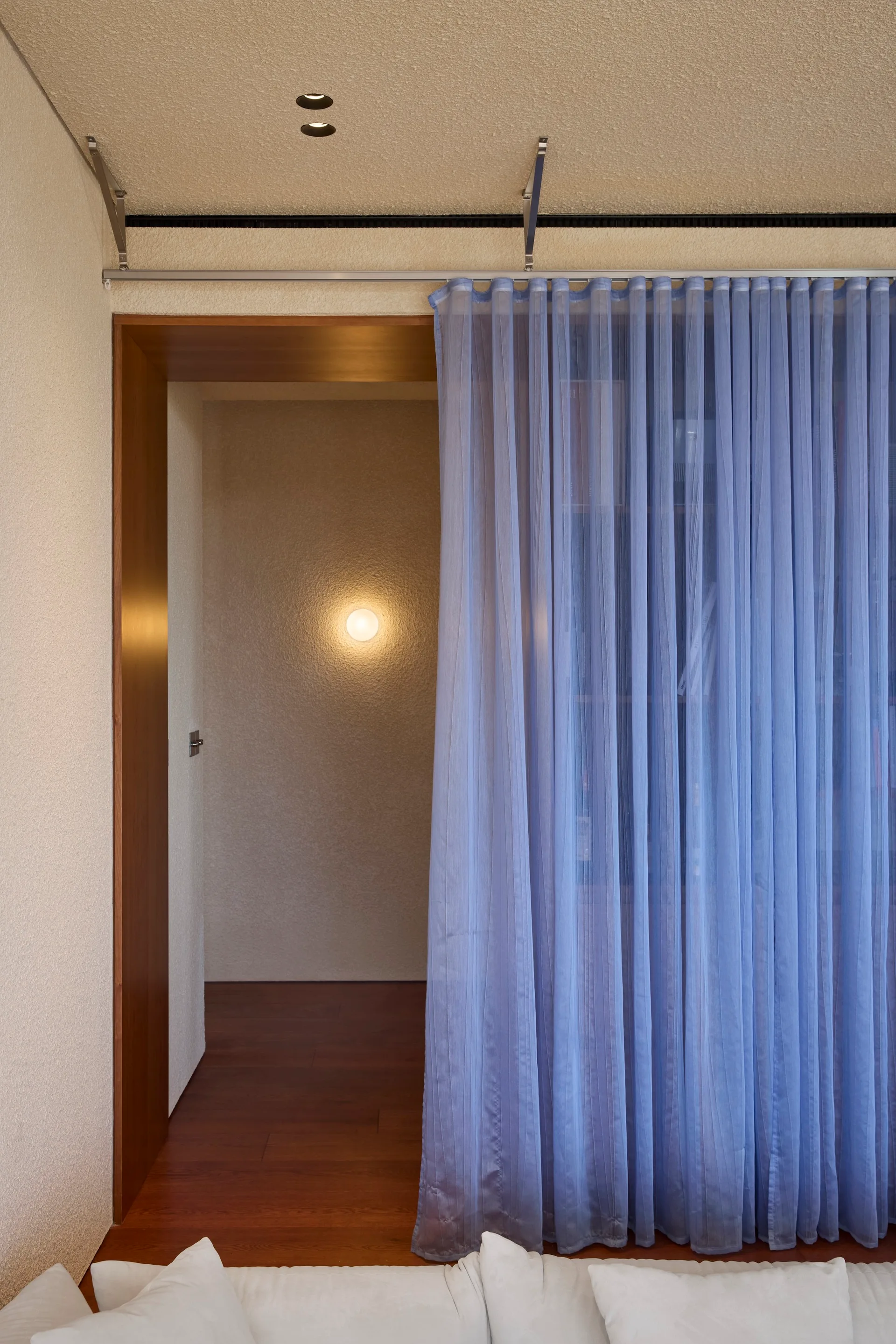 A wood-framed entryway features a sheer blue curtain hanging from a ceiling track next to textured cream walls and a glowing wall sconce.