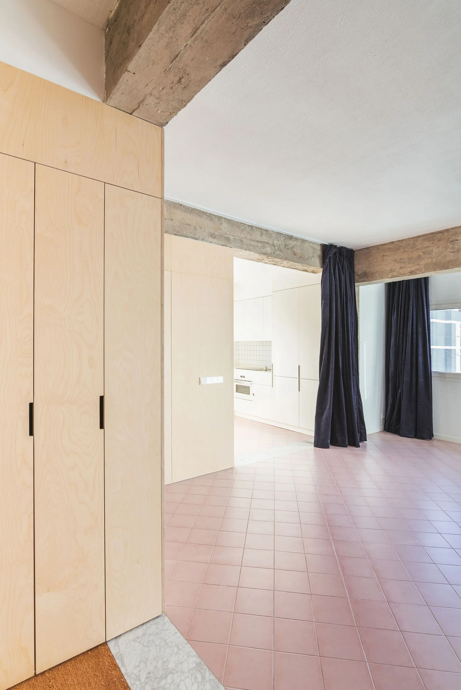 A minimalist interior featuring pink square tile flooring, built-in light wood cabinetry, and raw exposed concrete ceiling beams.