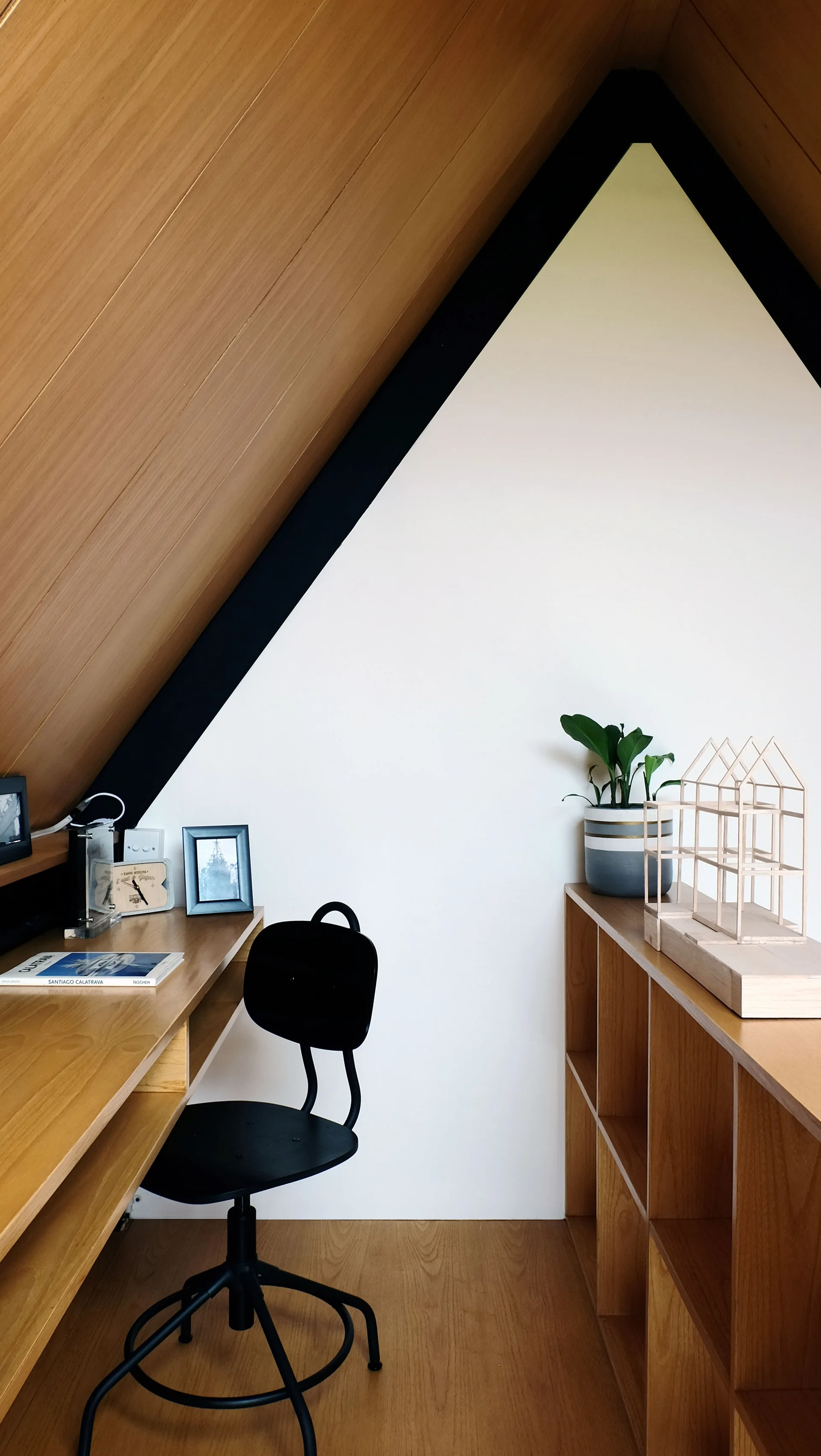 A compact home office in an attic nook featuring a built-in wooden desk, a black swivel chair, and open wooden shelving under a sloped plank ceiling.