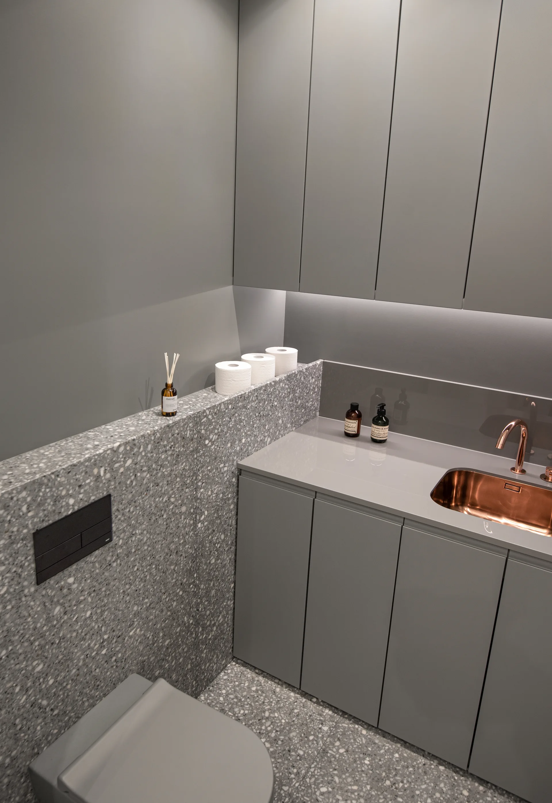 A minimalist grey powder room features a matching terrazzo floor and ledge, matte grey cabinets, and a polished copper sink with a matching faucet.
