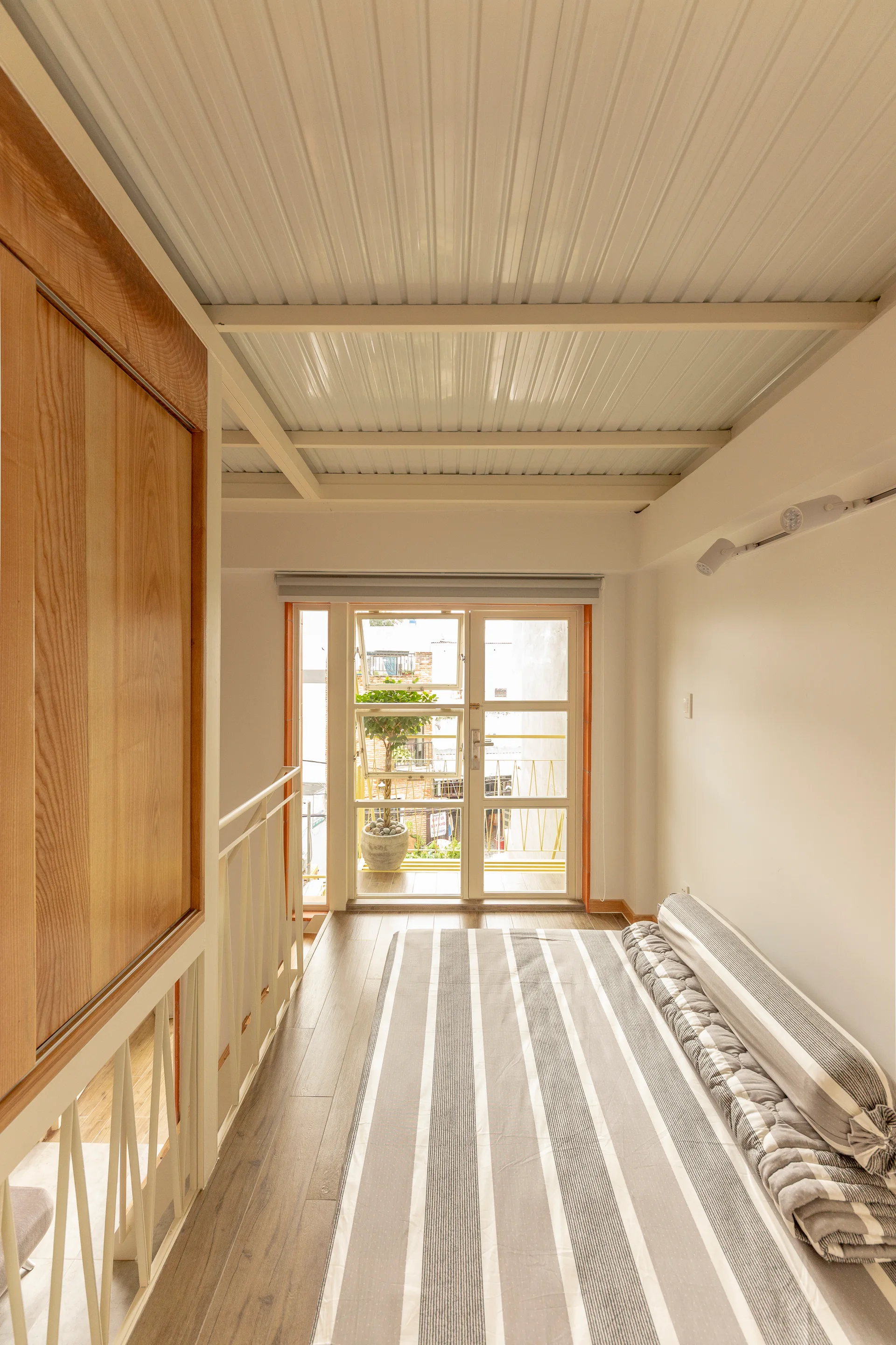 A compact loft bedroom features a striped mattress on the wooden floor, a large wooden closet door, and glass doors leading to a balcony.