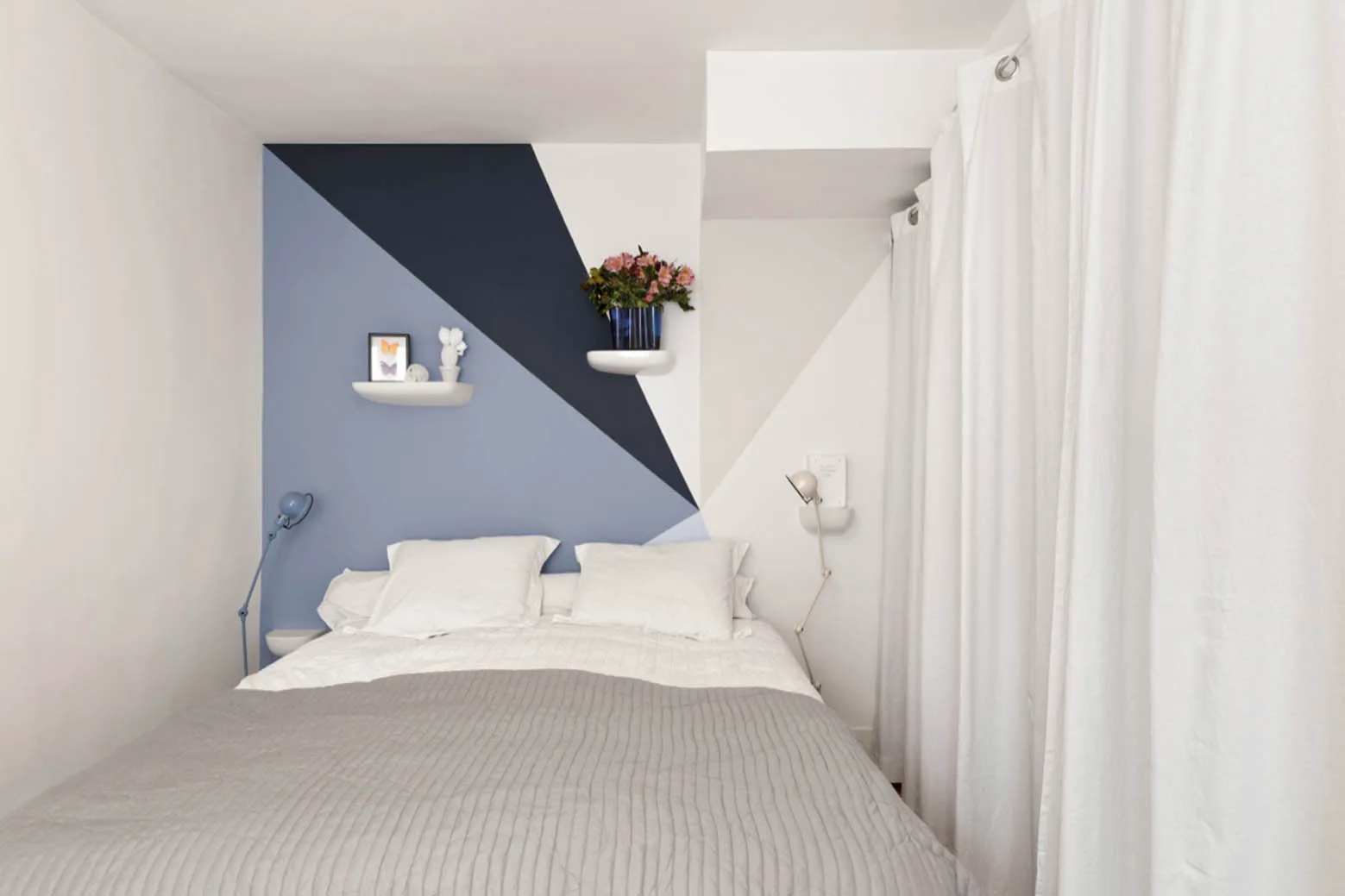 A small bedroom features a bed with white pillows and a grey blanket set against a blue and white geometric painted wall.
