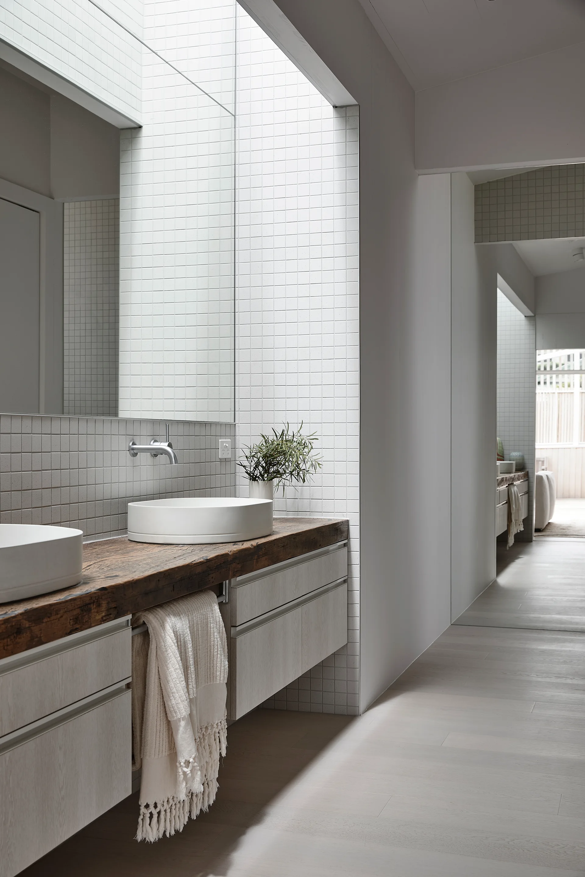 A minimalist bathroom features white vessel sinks on a thick reclaimed wood countertop set against a square-grid tiled wall.