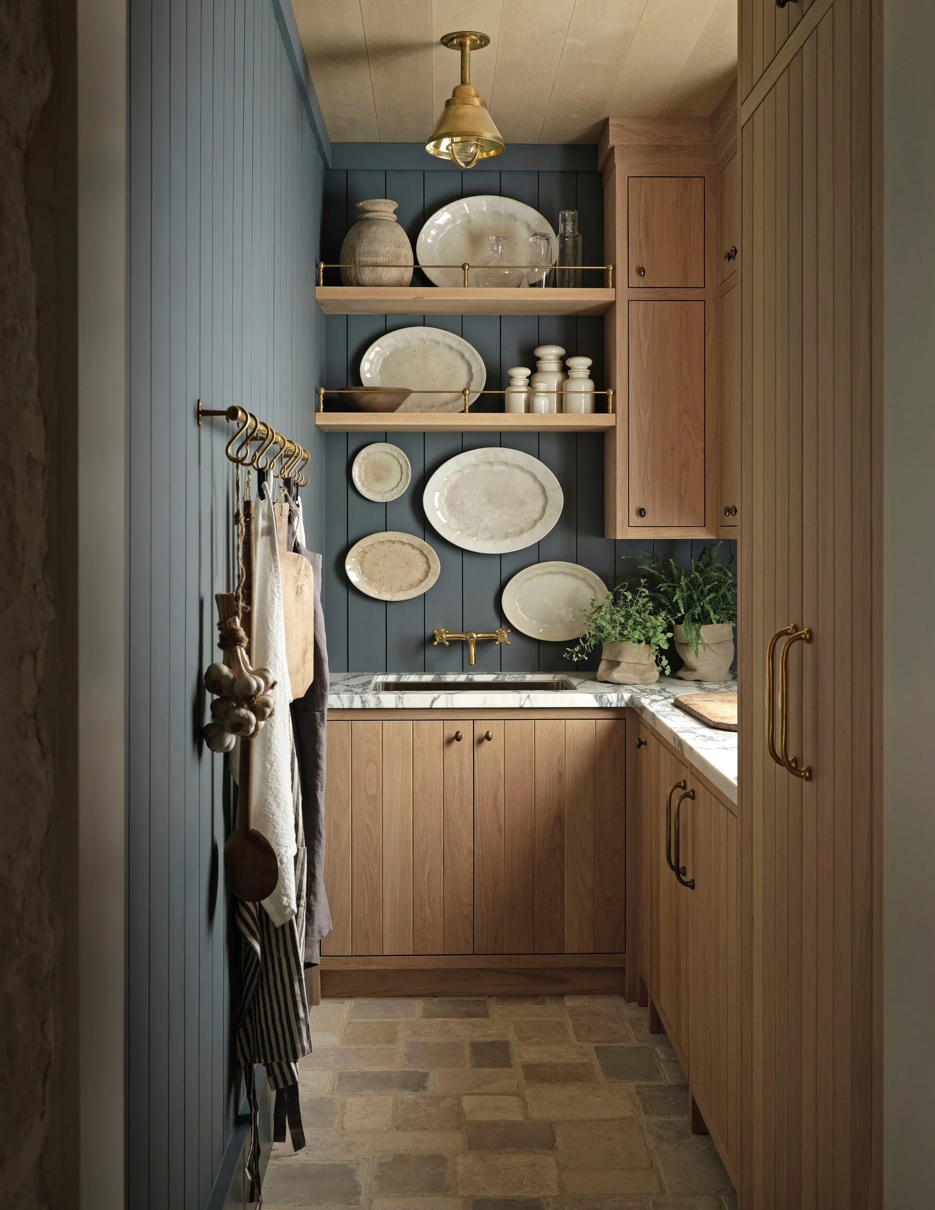 A narrow butler pantry features charcoal blue vertical wall paneling, natural oak cabinetry, marble countertops, and ceramic plates displayed on open shelves.