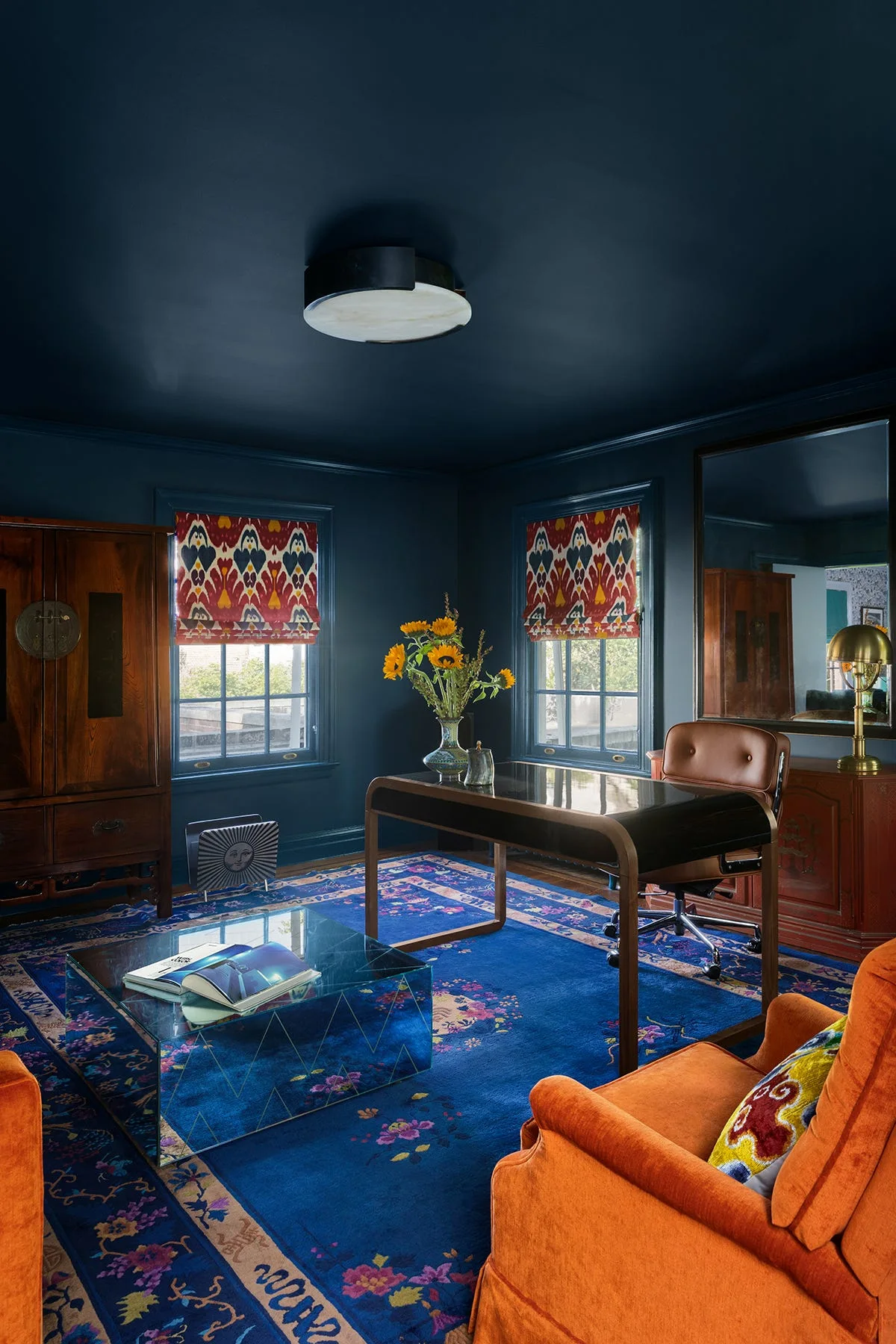 A home office features navy blue walls and ceiling, a dark wood desk, patterned roman shades, a blue rug, and orange armchairs.