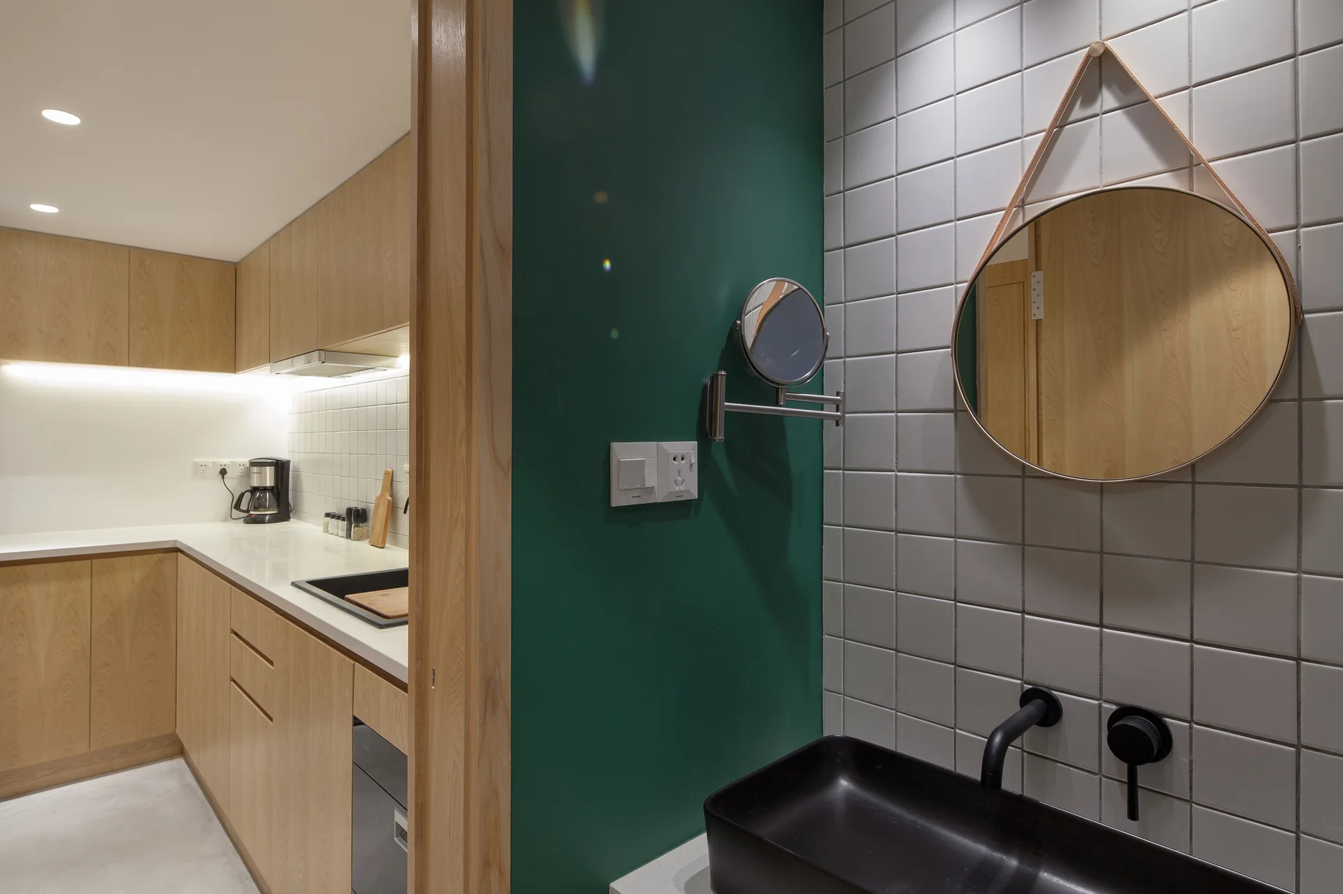 A bathroom vanity featuring a black vessel sink and round mirror on grey tiles, adjacent to a green wall and a wood-cabinet kitchen.