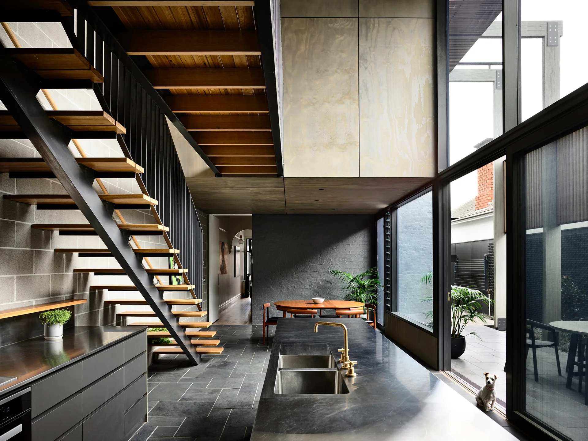 A modern kitchen features a dark stone island with a brass faucet, an open-riser wooden staircase, and large sliding glass doors.