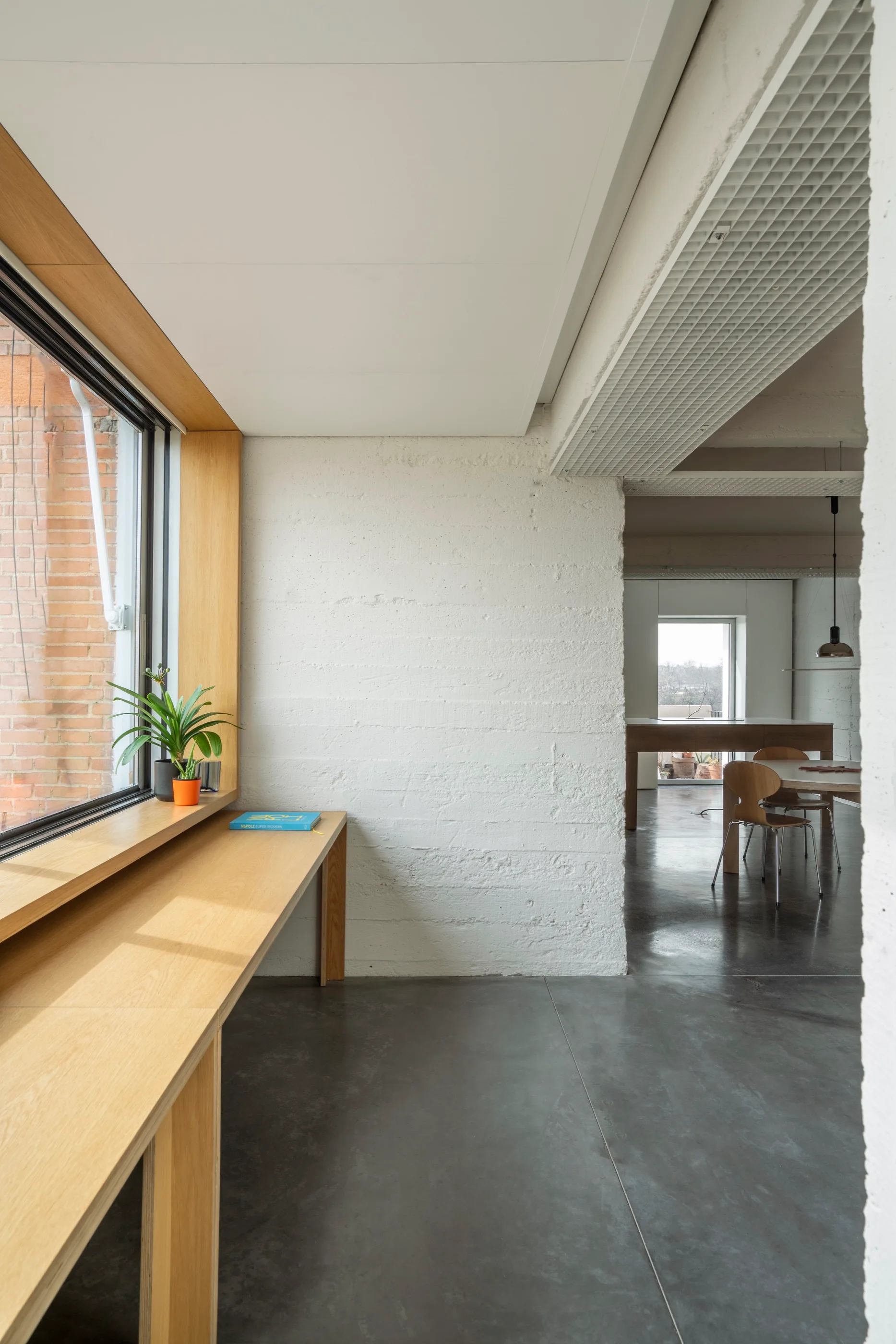 A long wood desk runs beneath a large window next to a textured white concrete wall and polished concrete floors leading to a dining area.