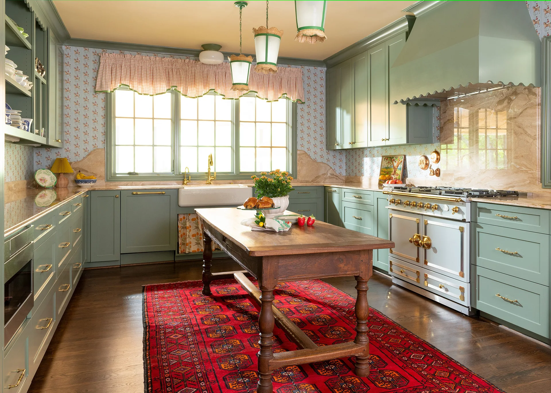 A traditional farmhouse kitchen features sage green cabinetry, a central wooden table island, and a vibrant red rug.