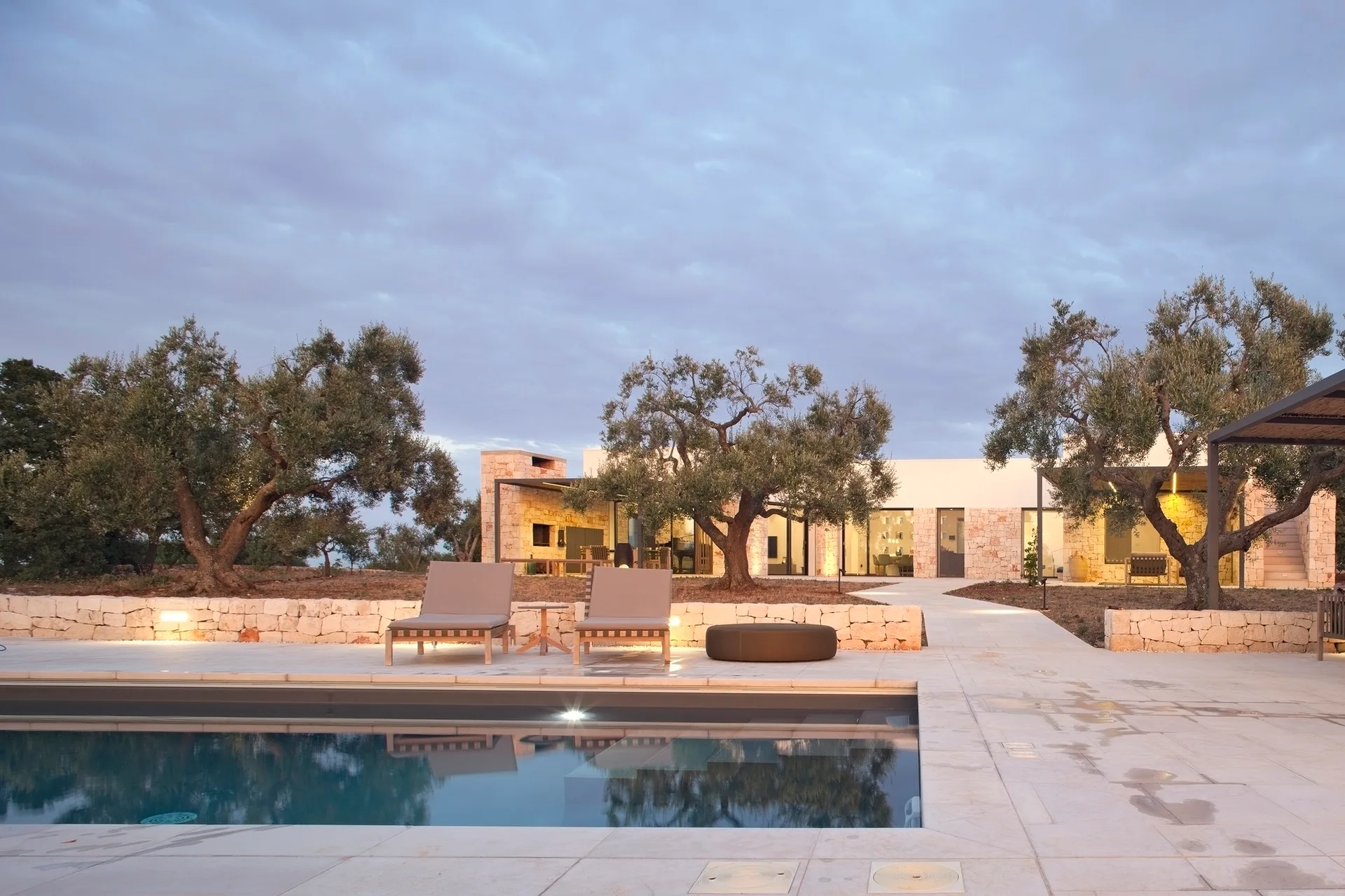A rectangular swimming pool sits next to a light-colored stone patio with two lounge chairs, framed by a low stone wall and mature olive trees at dusk.