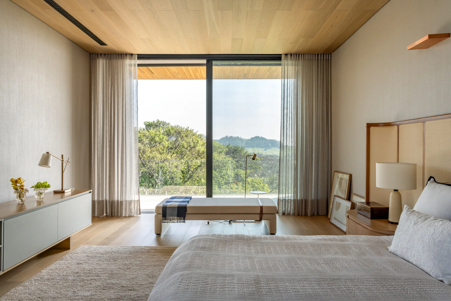 A minimalist bedroom featuring a bed with white bedding, an upholstered bench, sheer curtains, and a natural wood paneled ceiling.