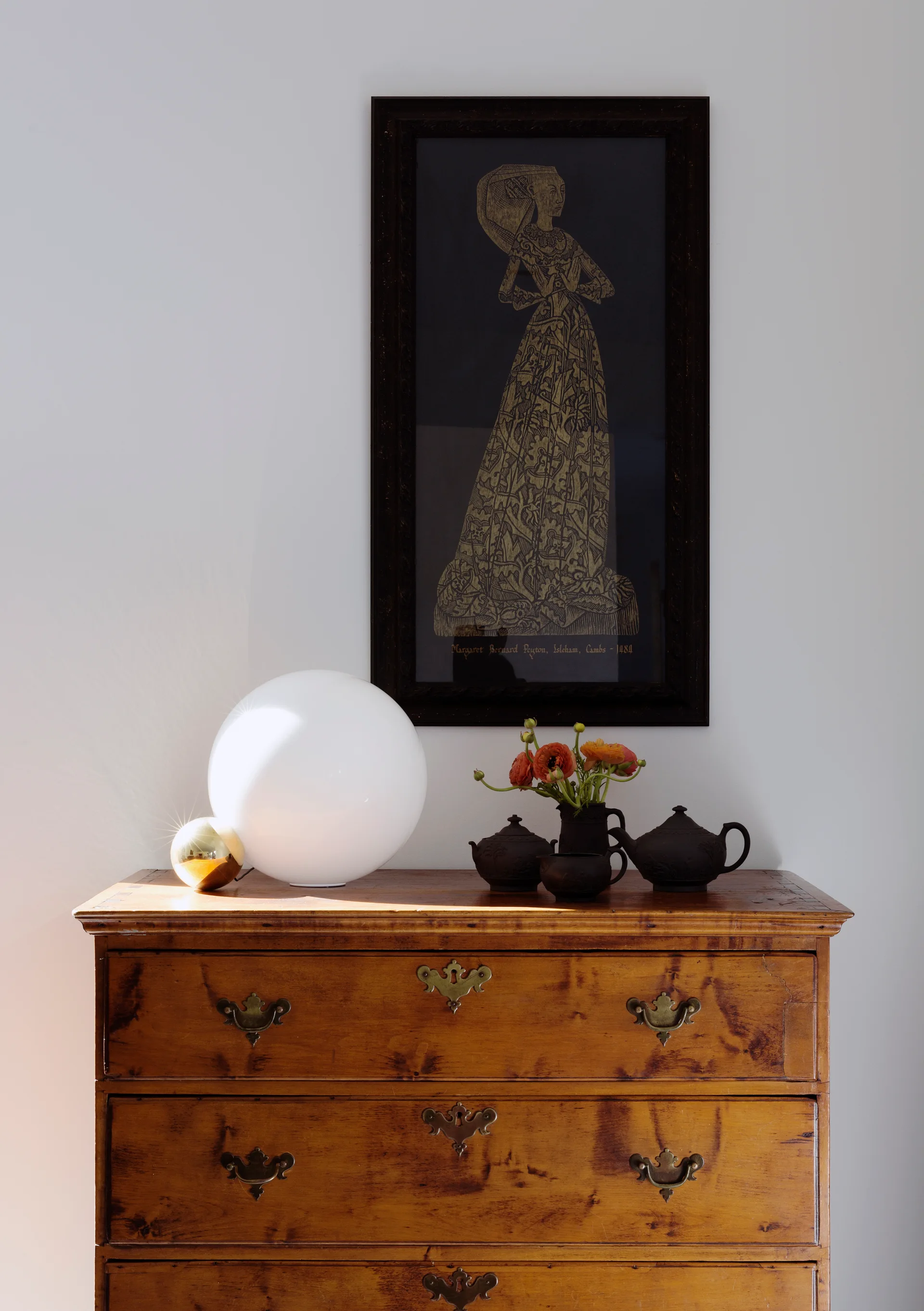 A warm wood antique chest of drawers styled with a glowing white globe lamp, dark ceramic tea set, and framed vertical artwork.