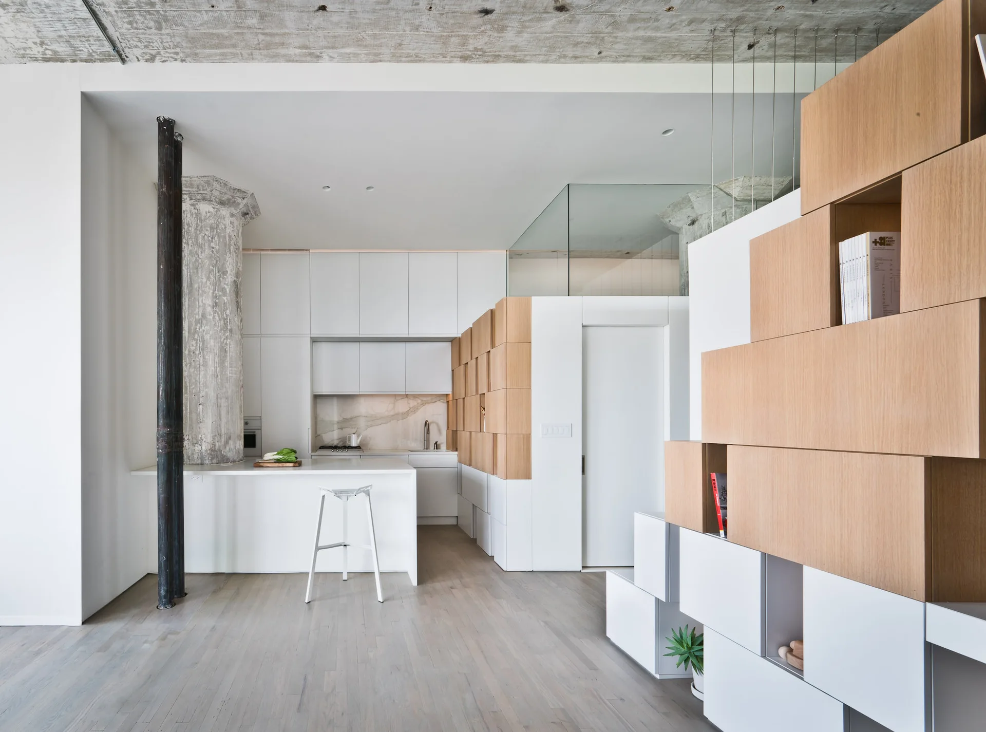 A modern loft kitchen featuring white flat-front cabinets, a raw concrete column and ceiling, and a modular wood and white block storage partition.