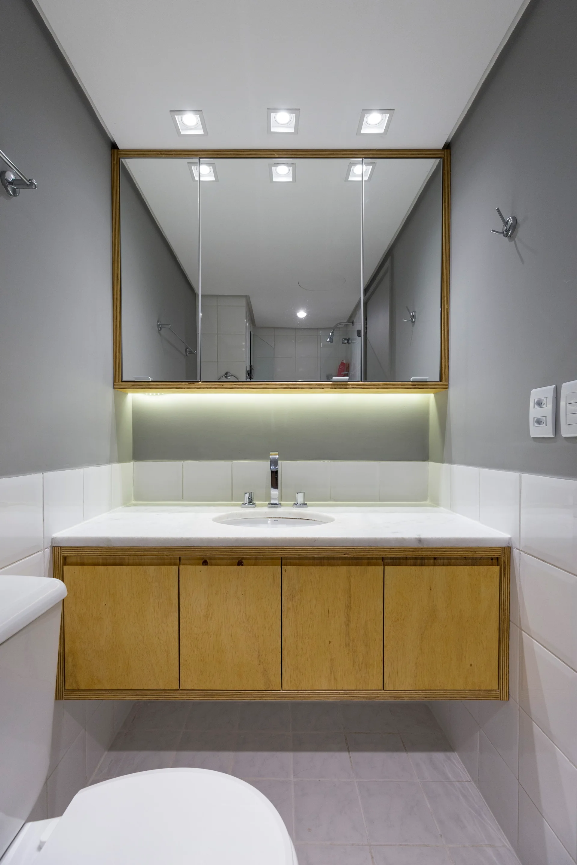 A modern bathroom features a floating wood vanity with a white countertop and sink below a large backlit mirrored cabinet against gray walls.