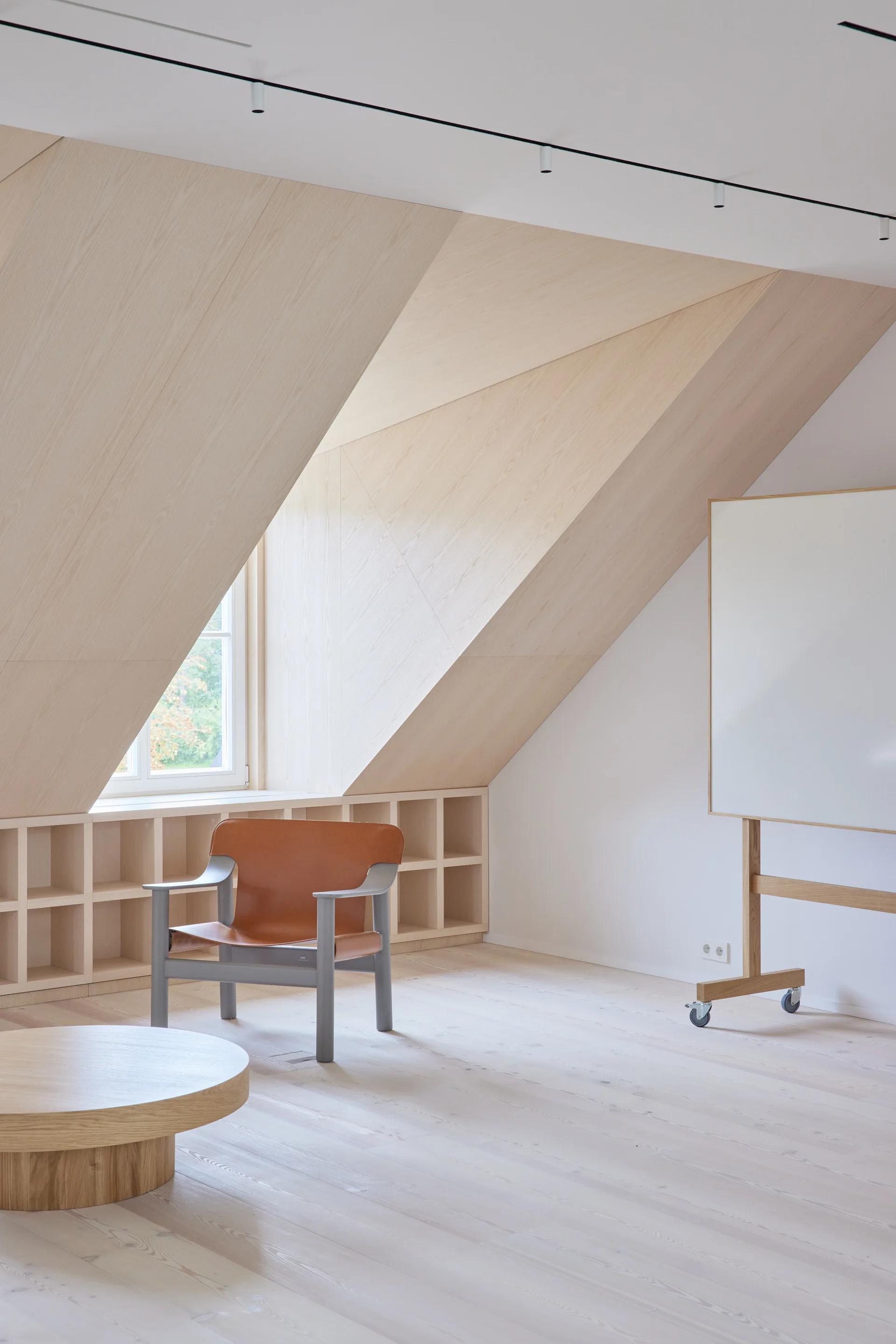A minimalist attic workspace features sloped light-wood paneling, a tan leather armchair, and a mobile whiteboard on a wooden stand.