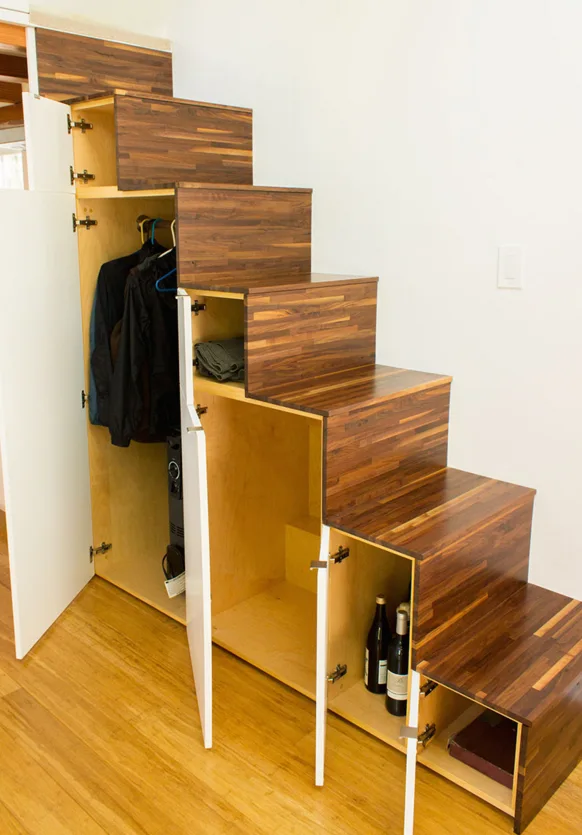 A custom wooden staircase unit features integrated storage compartments with open white cabinet doors revealing hanging jackets and bottles.