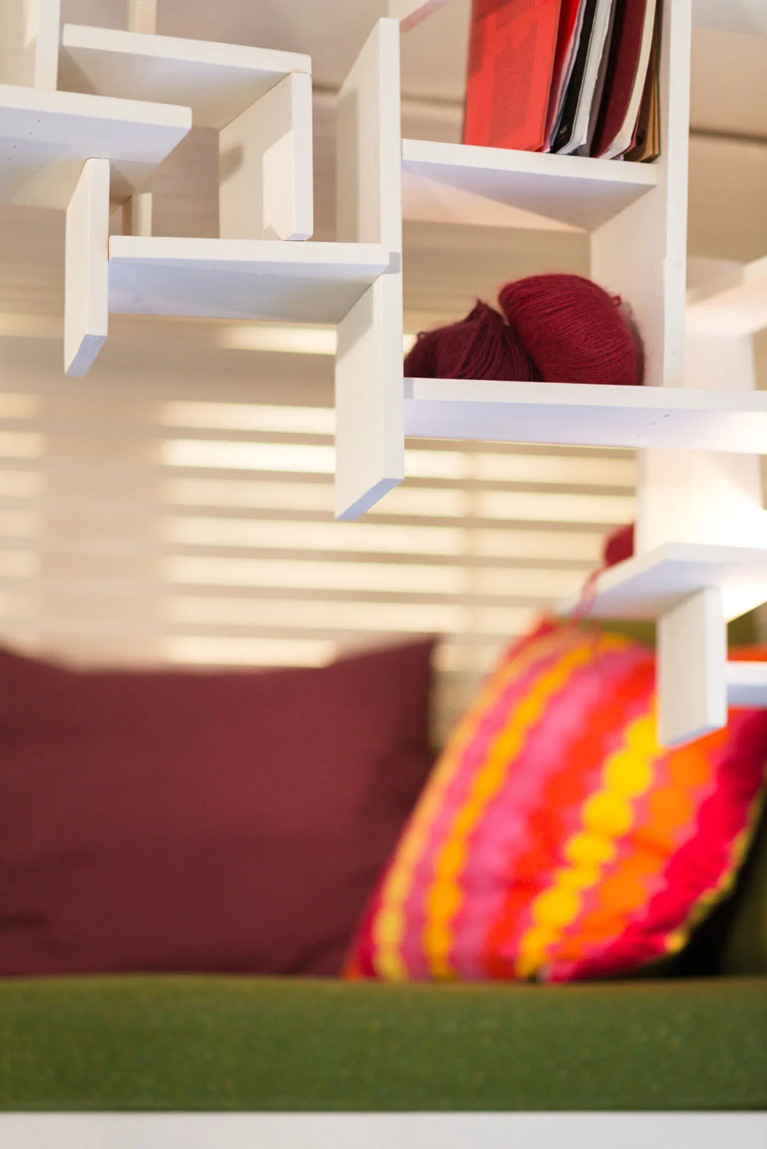A custom white geometric wooden shelving unit holds maroon yarn and books above a green sofa with colorful patterned pillows.