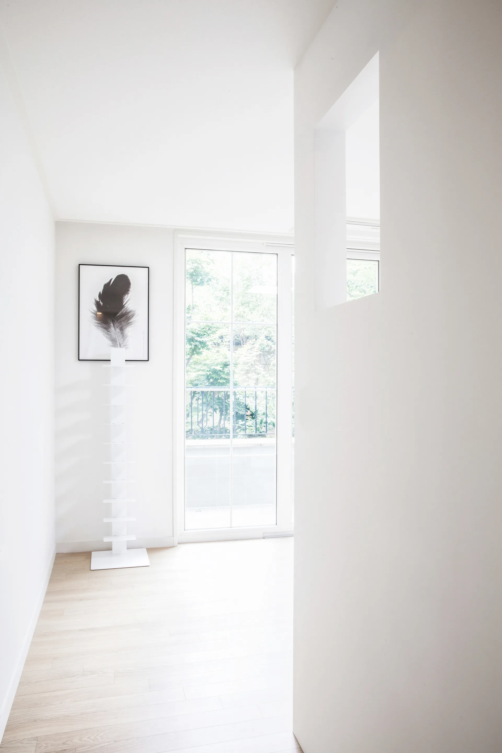 A bright minimalist hallway features light wood floors, white walls, a tall spine bookcase, and a framed print of a black feather.