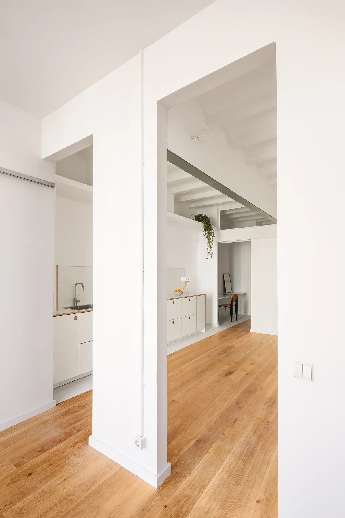 Minimalist interior with white partition walls, rectangular doorless openings, and light wood flooring leading to kitchen cabinetry.