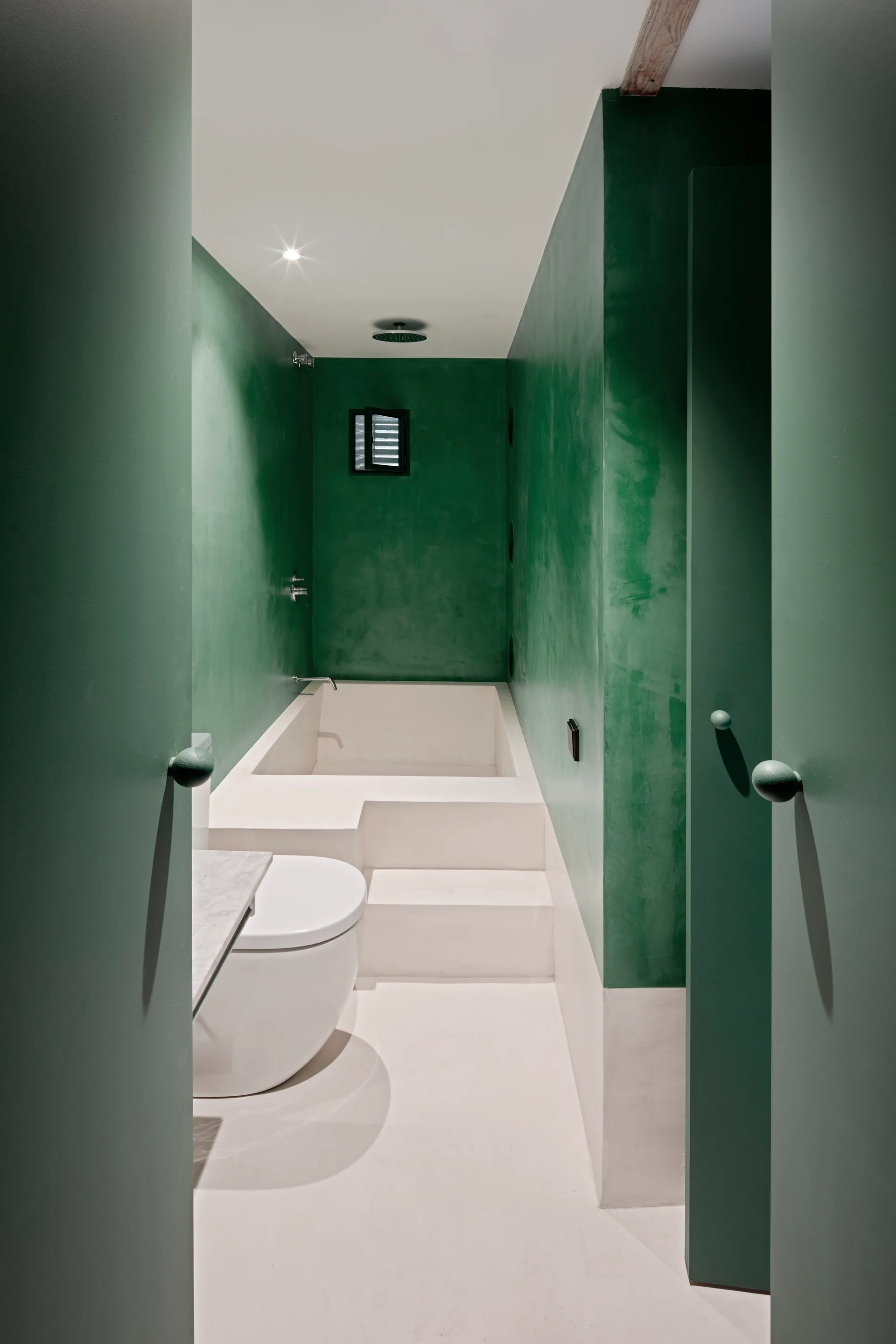 A minimalist bathroom with rich forest green plaster walls, a white floor, and a built-in sunken bathtub accessed by white concrete steps.