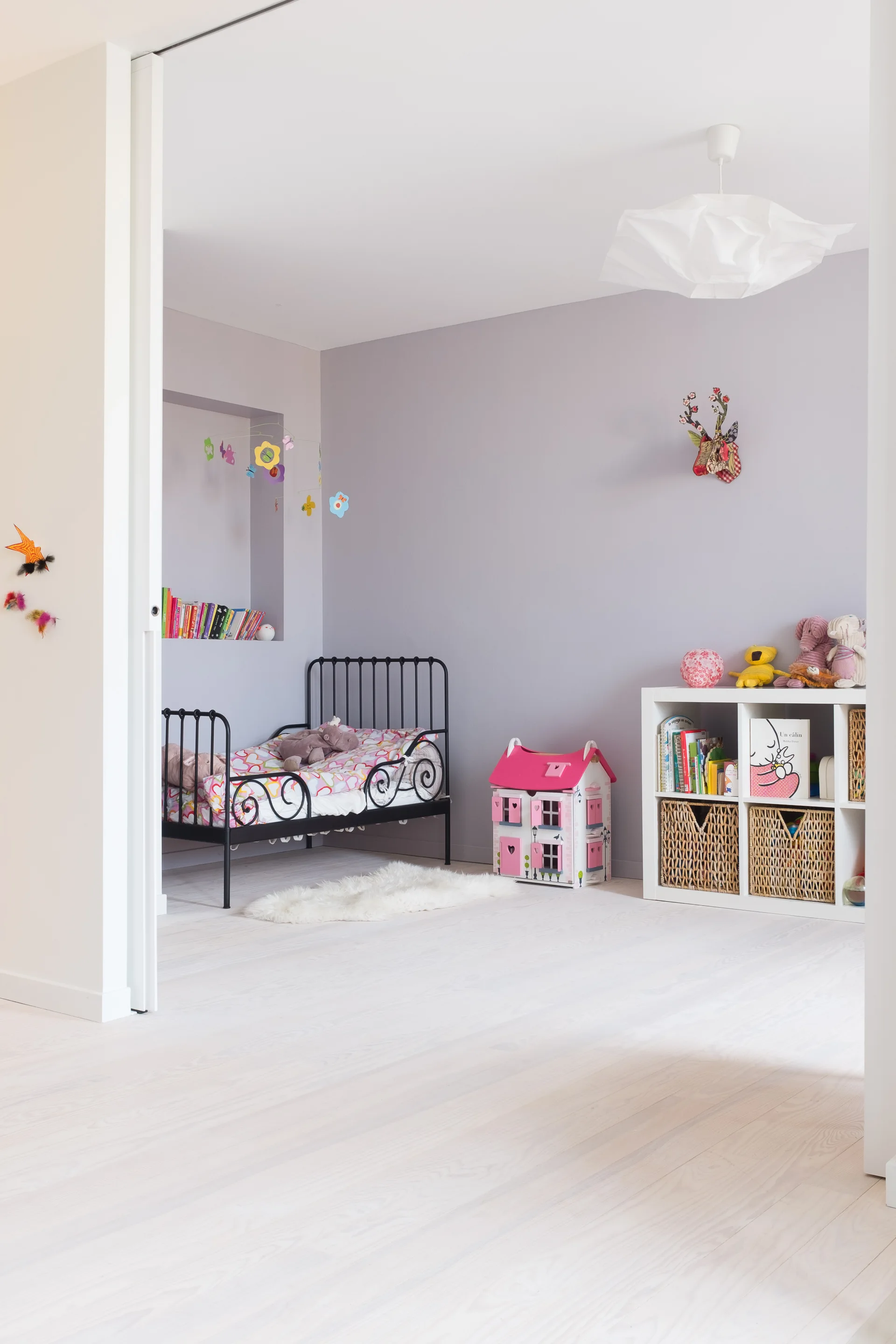A child's bedroom features a black metal scrollwork bed, lavender walls, and a white cube storage unit on white wood flooring.