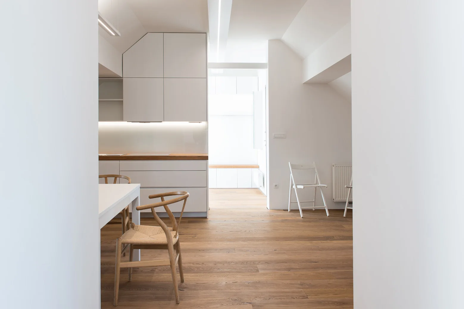 A minimalist kitchen features white handleless cabinets, a natural wood countertop, matching wood flooring, and a dining table with woven wood chairs.