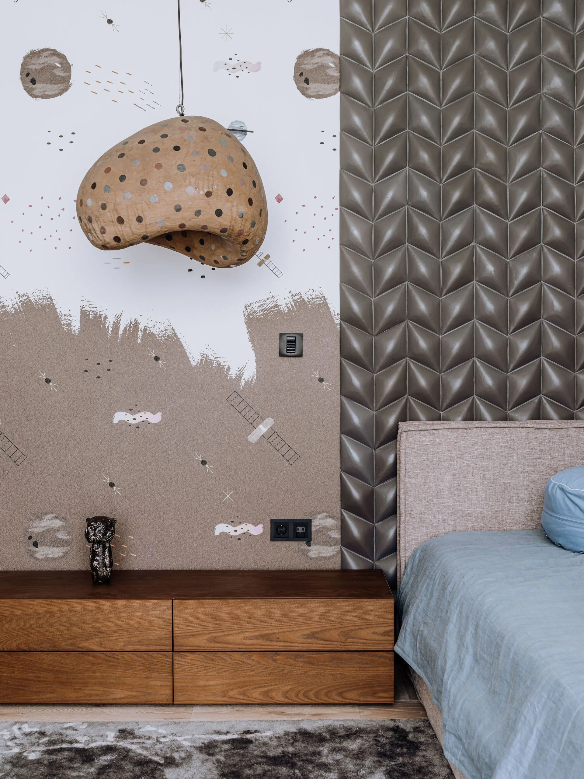 A bedroom detail showcasing a low wood nightstand, a fabric headboard, a polka-dotted pendant lamp, and a wall split with space-themed wallpaper and 3D tiles.
