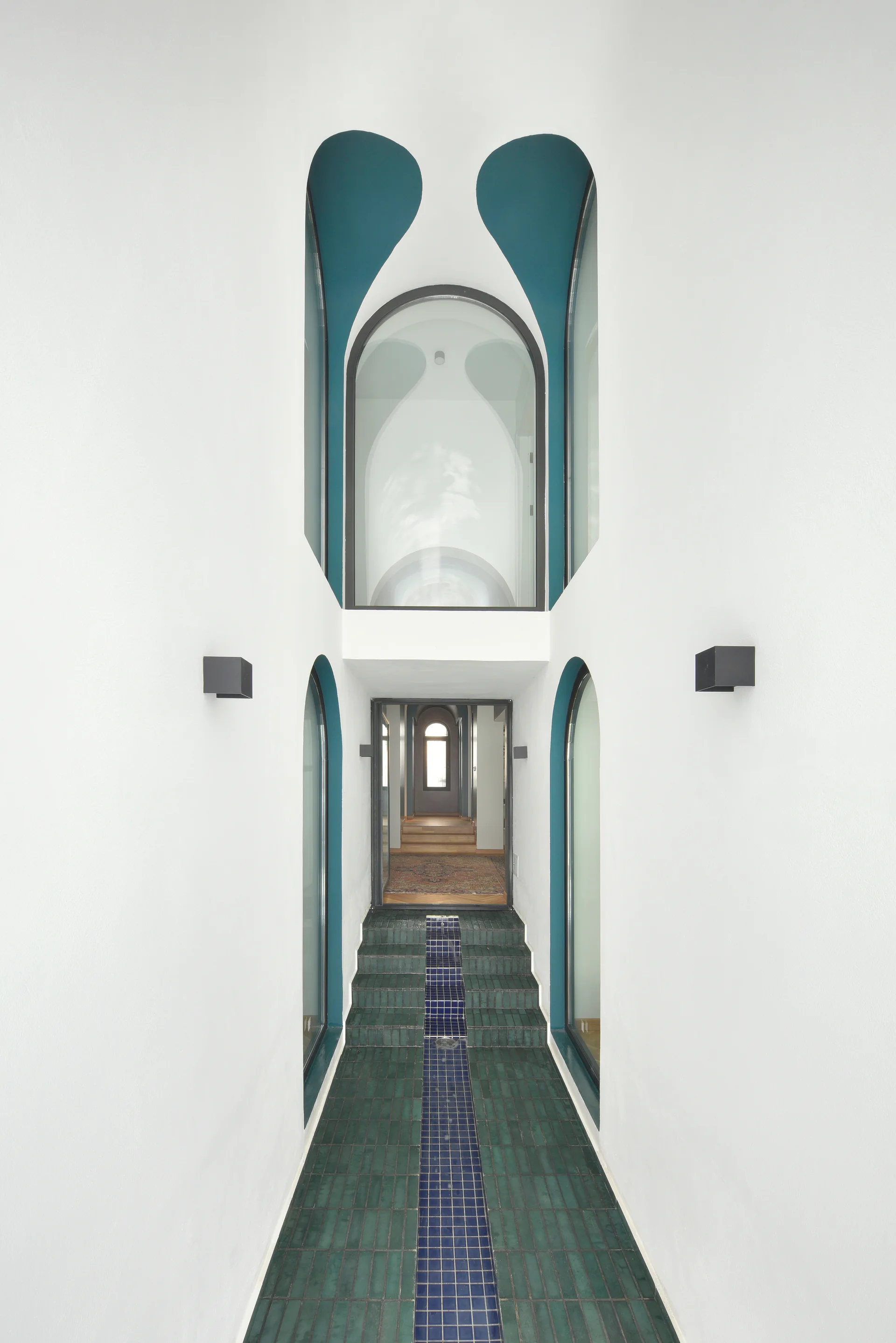A symmetrical white hallway features teal-painted arched window cutouts, modern black wall sconces, and a staircase tiled in dark green and blue.