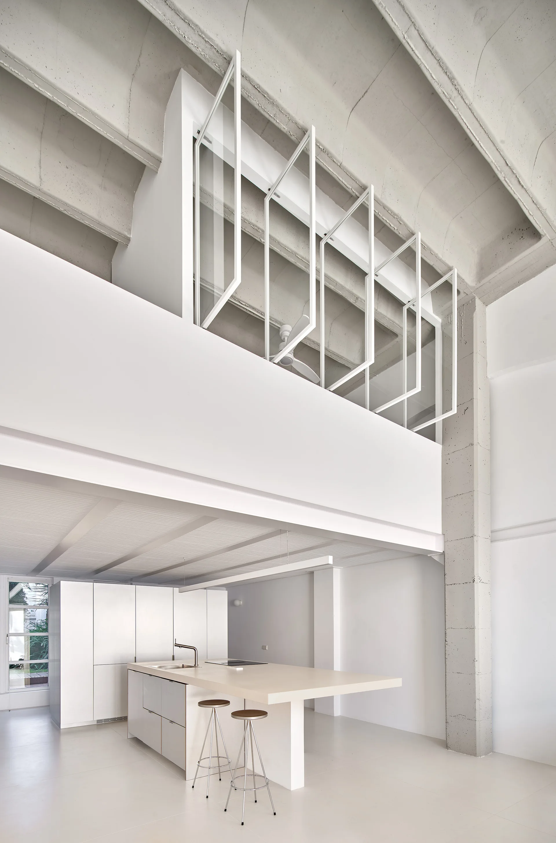 A minimalist white loft kitchen features a central island with stools beneath an exposed concrete ceiling and a mezzanine level with metal frames.