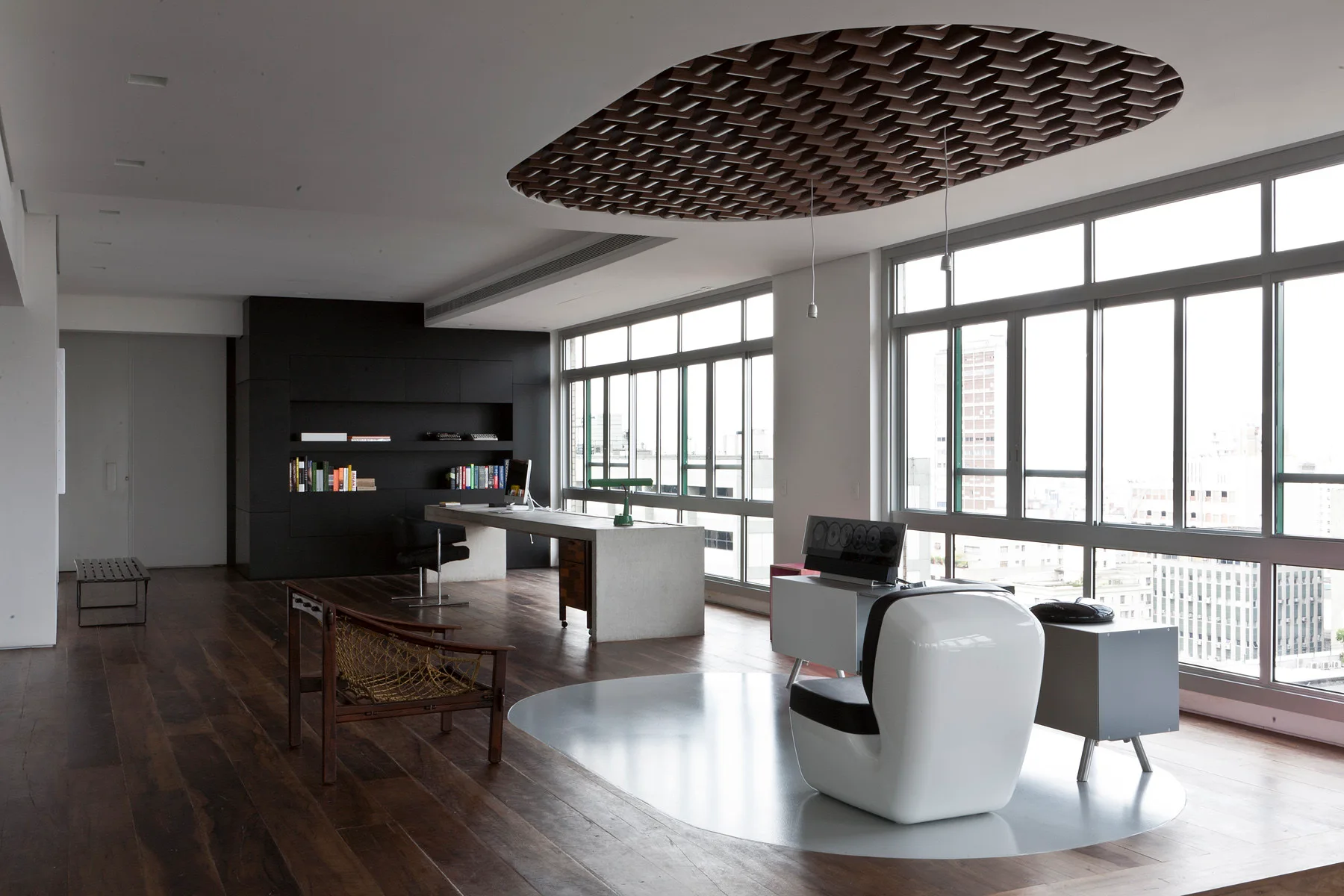 A spacious modern home office featuring a concrete desk, dark wood flooring, a sculptural white armchair, and an oval wooden ceiling cutout.
