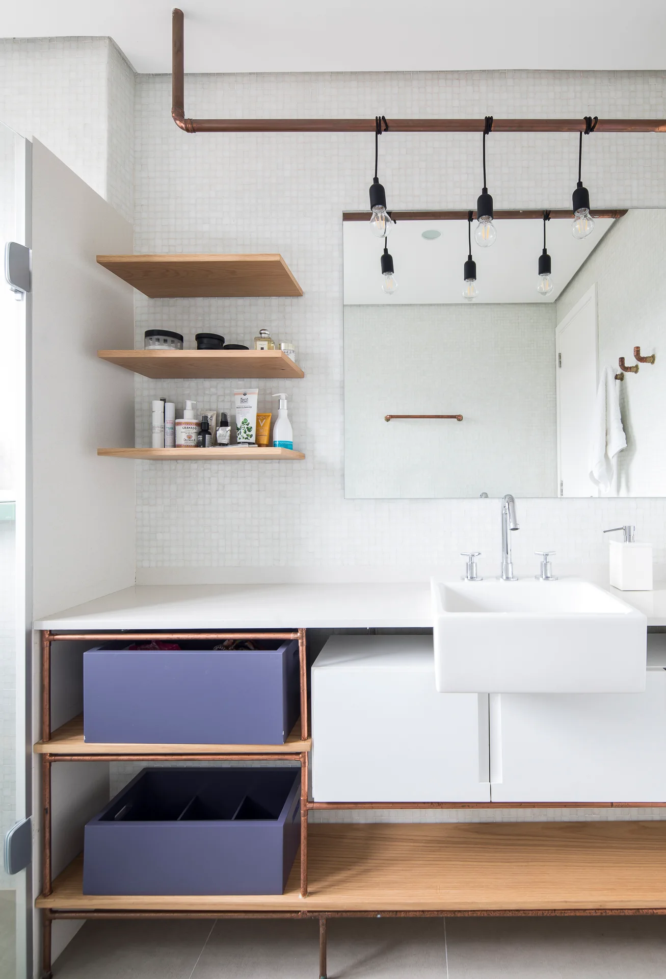 A modern bathroom vanity featuring a white sink, exposed copper pipe detailing, floating wooden shelves, and purple storage bins.