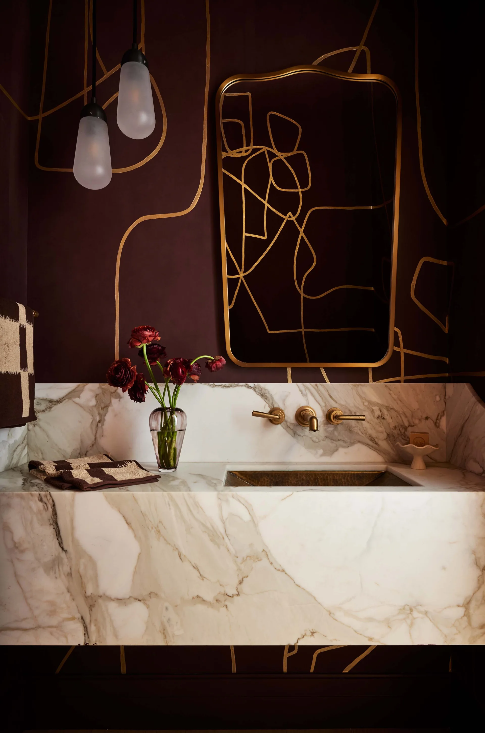 A powder room features a heavily veined marble vanity, deep burgundy walls with abstract gold lines, a brass mirror, and a wall-mounted brass faucet.